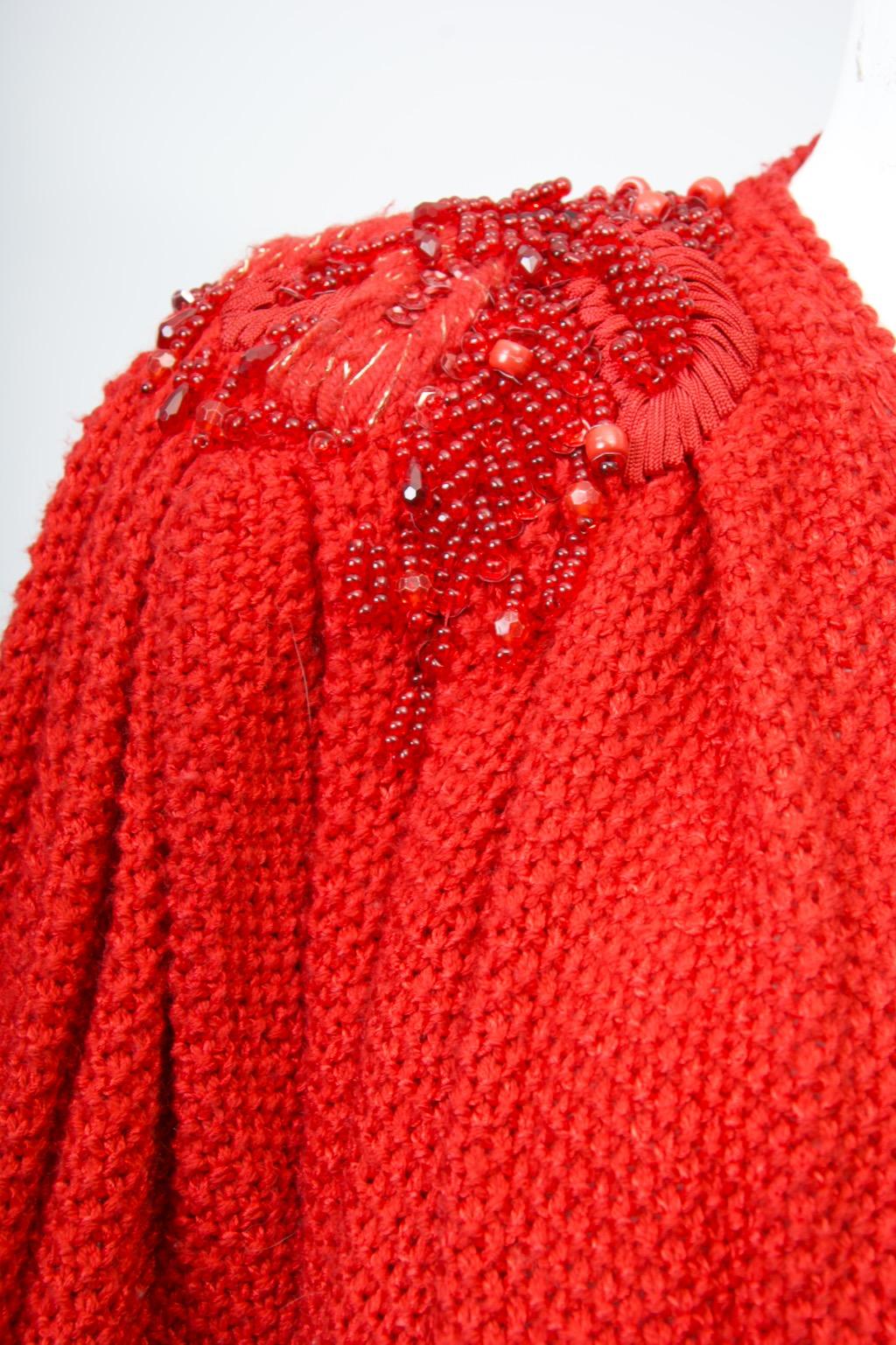 1980s red knit cardigan elaborately embellished at the shoulders with appliquéd yarn, beads, and sequins. The sweater features an oversized cut, a deep V neckline with coordinating buttons, and full sleeves that are pleated from the shoulders. The