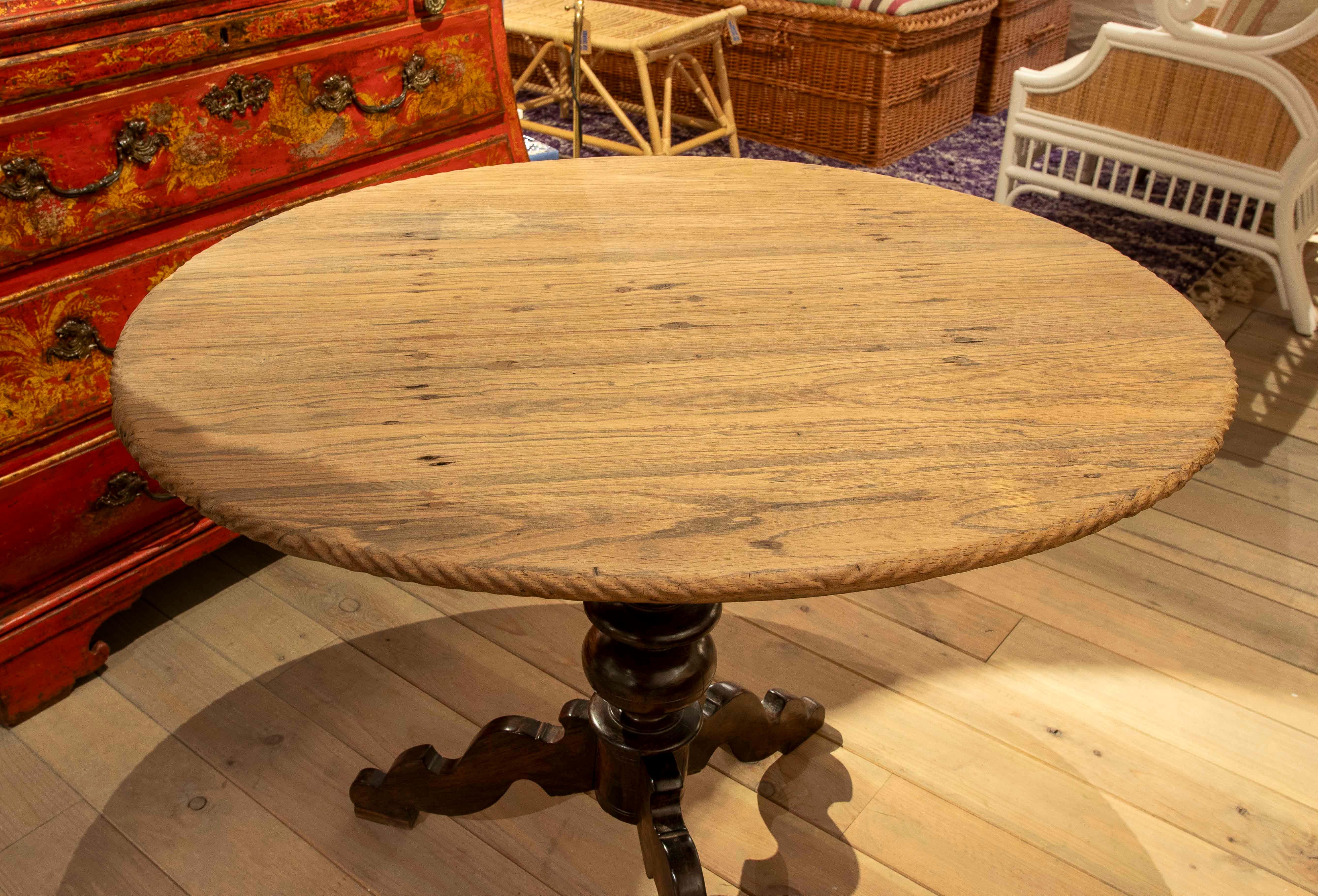 Round dining table with a light wood top in a natural finish that highlights the grain and texture of the material. Its central base contrasts with an elegant turned dark wood base with three curved supports, lending a classic and robust feel to the