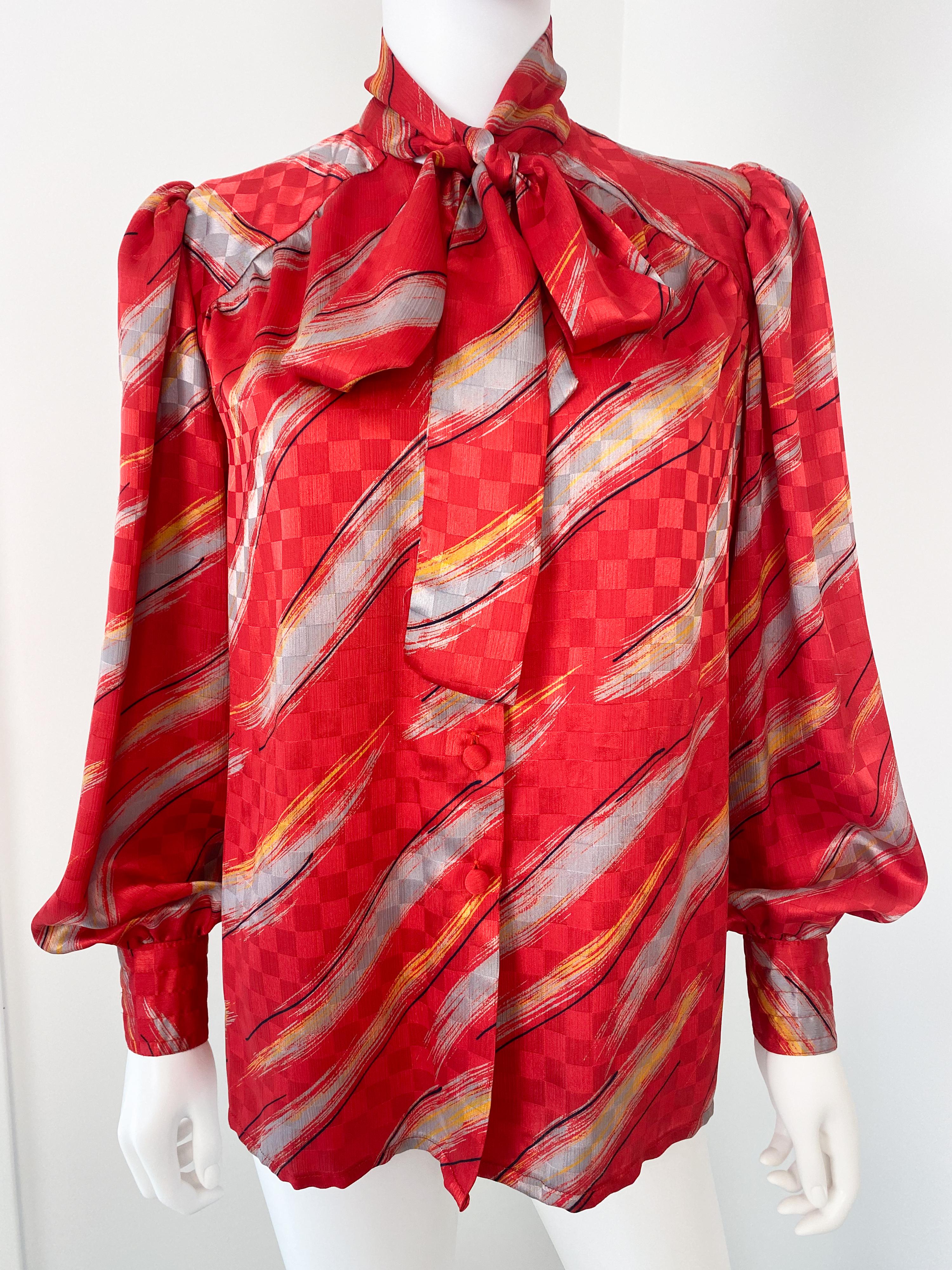 Cette blouse vintage des années 1980 est confectionnée en polyester-viscose soyeux et présente un élégant nœud papillon avec une écharpe attachée. Le tissu rouge vif est orné d'un motif audacieux en gris et jaune, accentué par un contraste noir pour
