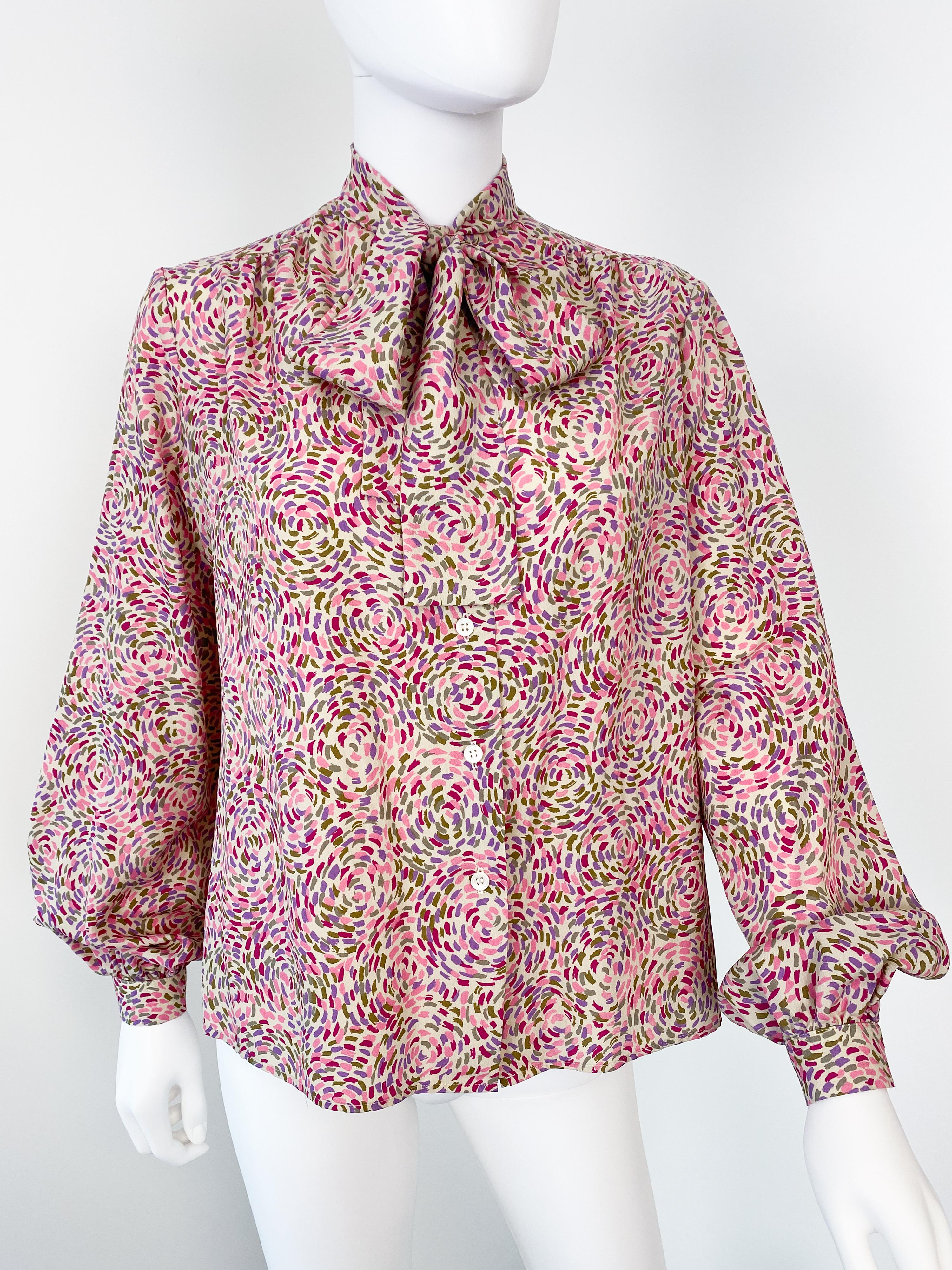 This exquisite vintage 1980s blouse is crafted from silky polyester viscose and features a classic pussy bow with its original tie. The elegant pointillism print showcases shades of pink, purple, burgundy, and green on an off-white background,