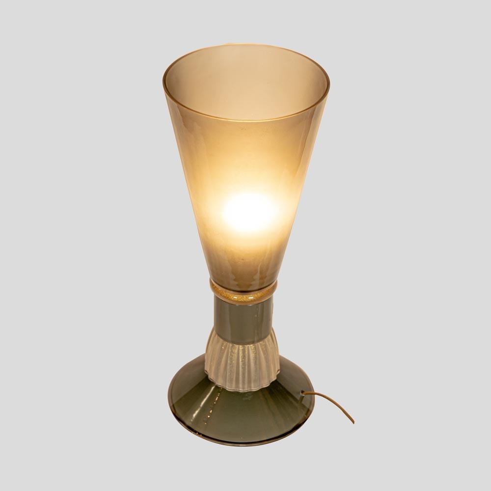 A very elegant table light shaped as a truncated cone on a circular base. Blown fume’ grey and clear with gold inclusions blown glass with decorative parts made in Murano, Venice, Italy in the 1980’s.
This particularly attractive table light has