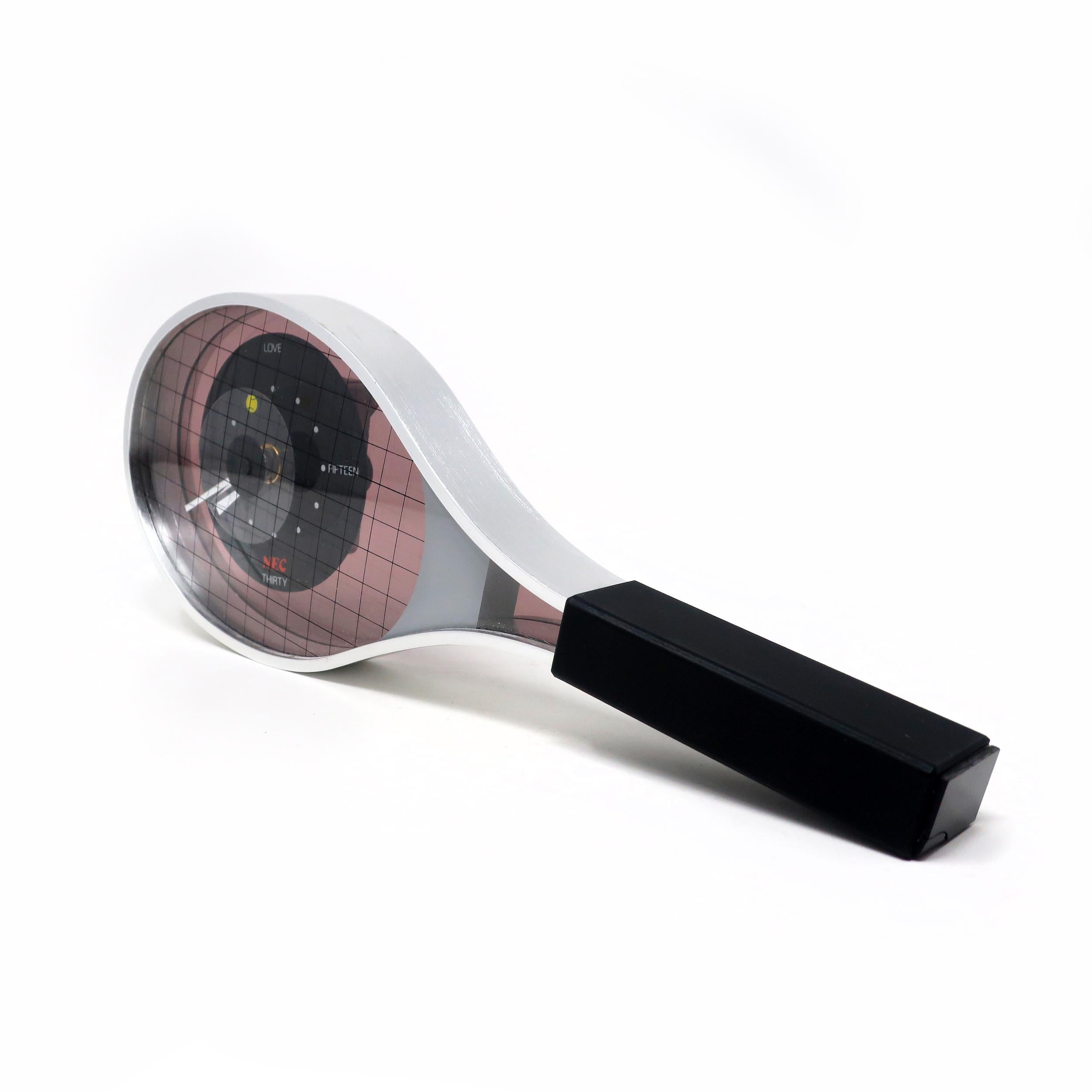 A whimsical desk or shelf clock in the shape of a tennis racket. It has a metal frame with a black plastic handle, a face that resembles the interwoven strings of a racket head, number marks like the scores in tennis (Love = 12, Fifteen = 3, Thirty