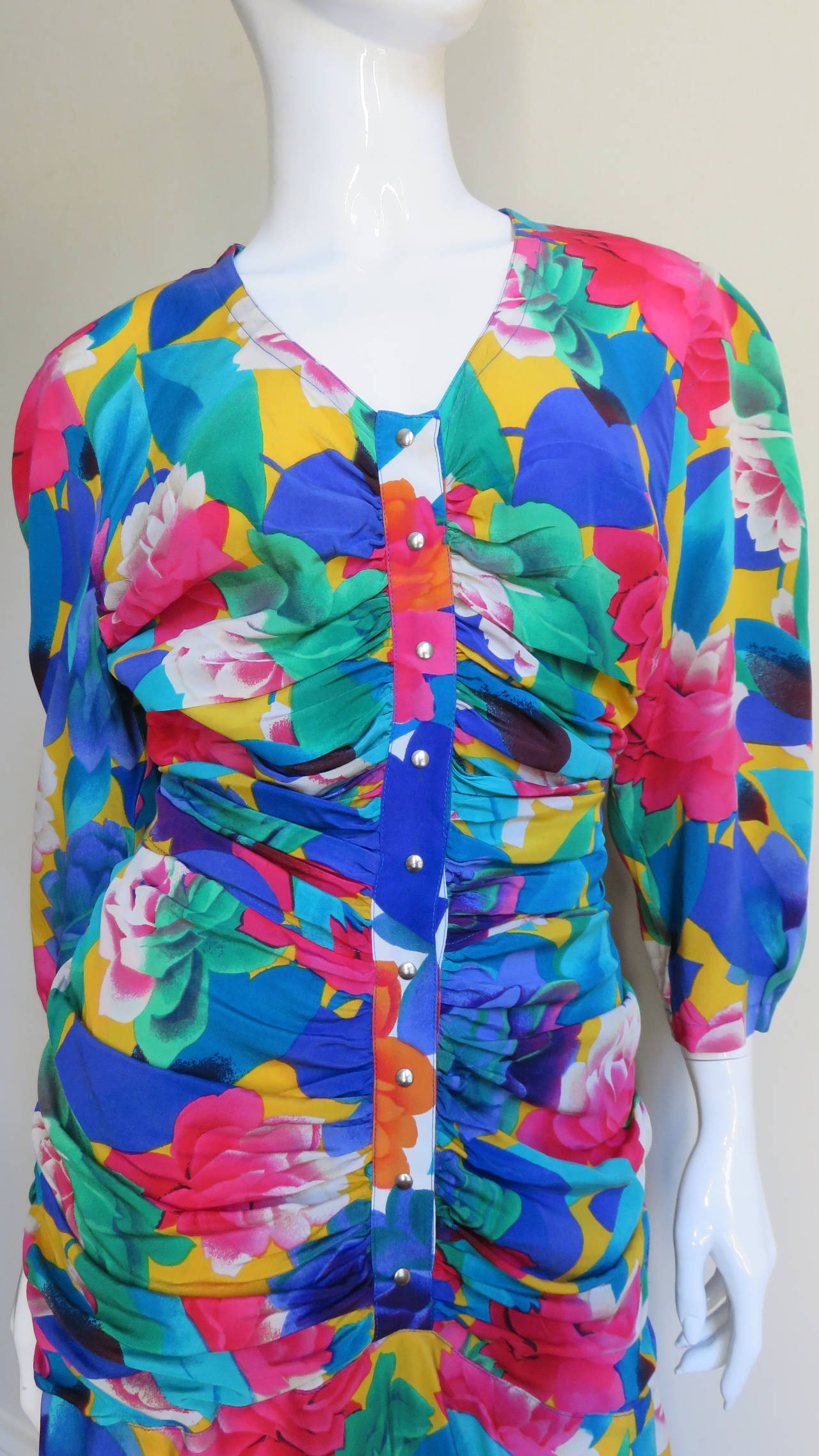 A brightly colored flower pattern silk dress from Thierry Mugler in blues, greens, pinks and yellow.  It has a V neck, 3/4 length sleeves and snaps along the center front of the horizontally ruched bodice.  It has is a back half belt with a blue