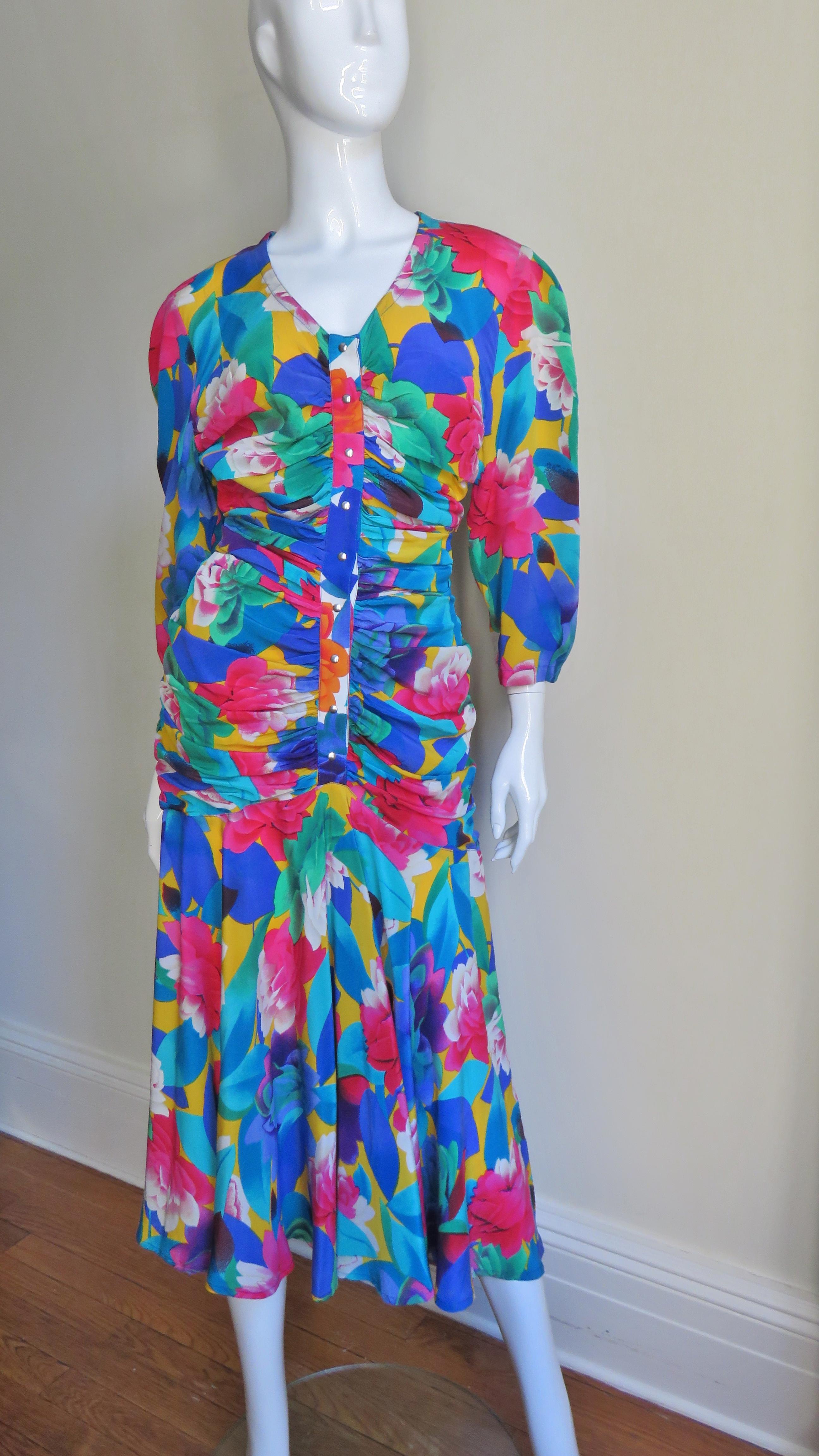 A brightly colored flower pattern silk dress from Thierry Mugler in blues, greens, pinks and yellow.  It has a V neck, 3/4 length sleeves and snaps along the center front of the ruched bodice.  There is a ruched cummerbund at the waist which wraps