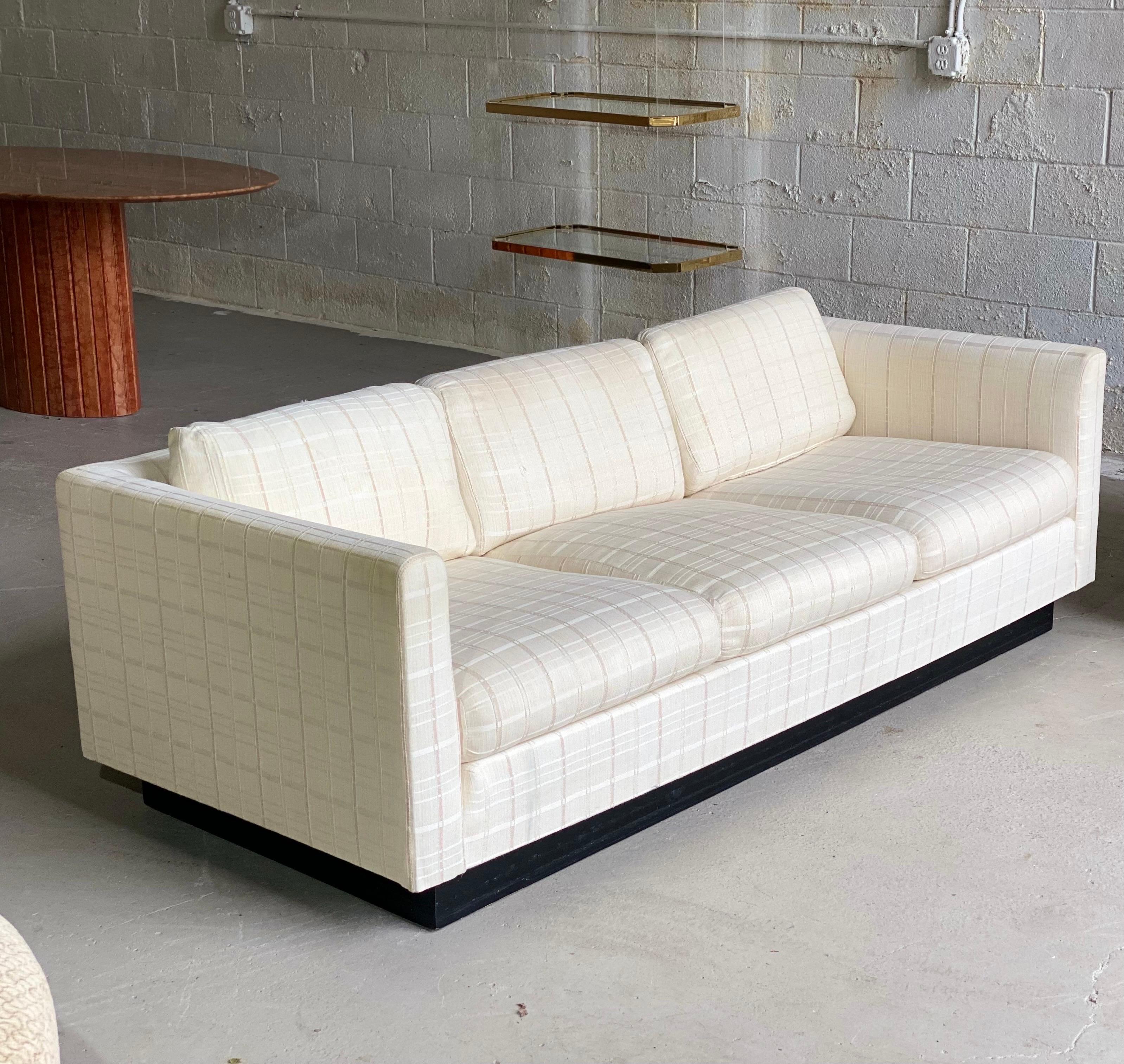 We are very pleased to offer a chic, tuxedo sofa in the style of Milo Baughman, circa the 1980s. This piece showcases a wraparound frame upholstered in a neutral linen fabric supported by a black plinth base. Some cushions have a few stains, but