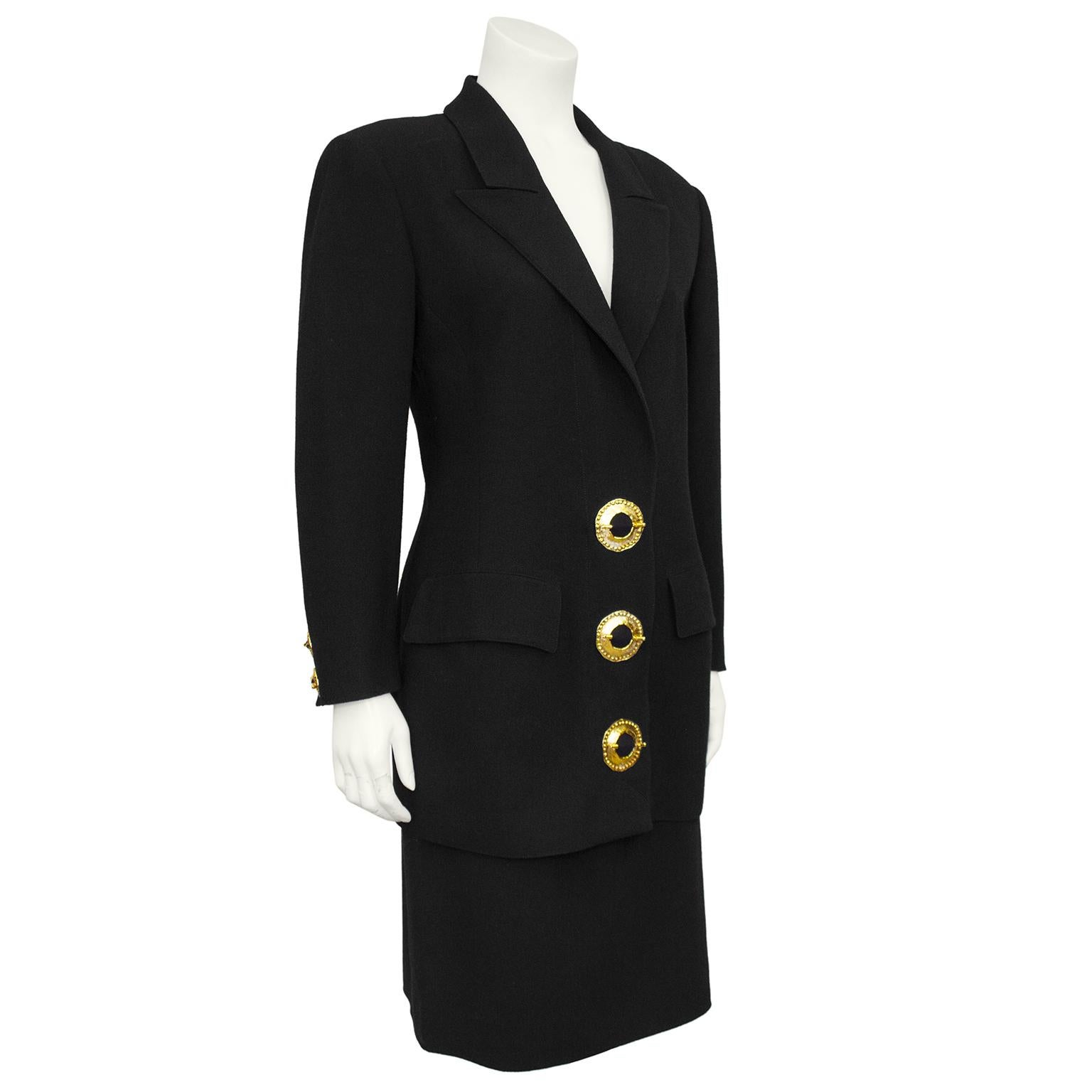 Valentino black wool skirt suit from the 1980s. Elongated blazer features large shoulder pads, notched collar and horizontal flap pockets. The black is contrasted with gold tone metal large circular buttons that look like brooches with pins through