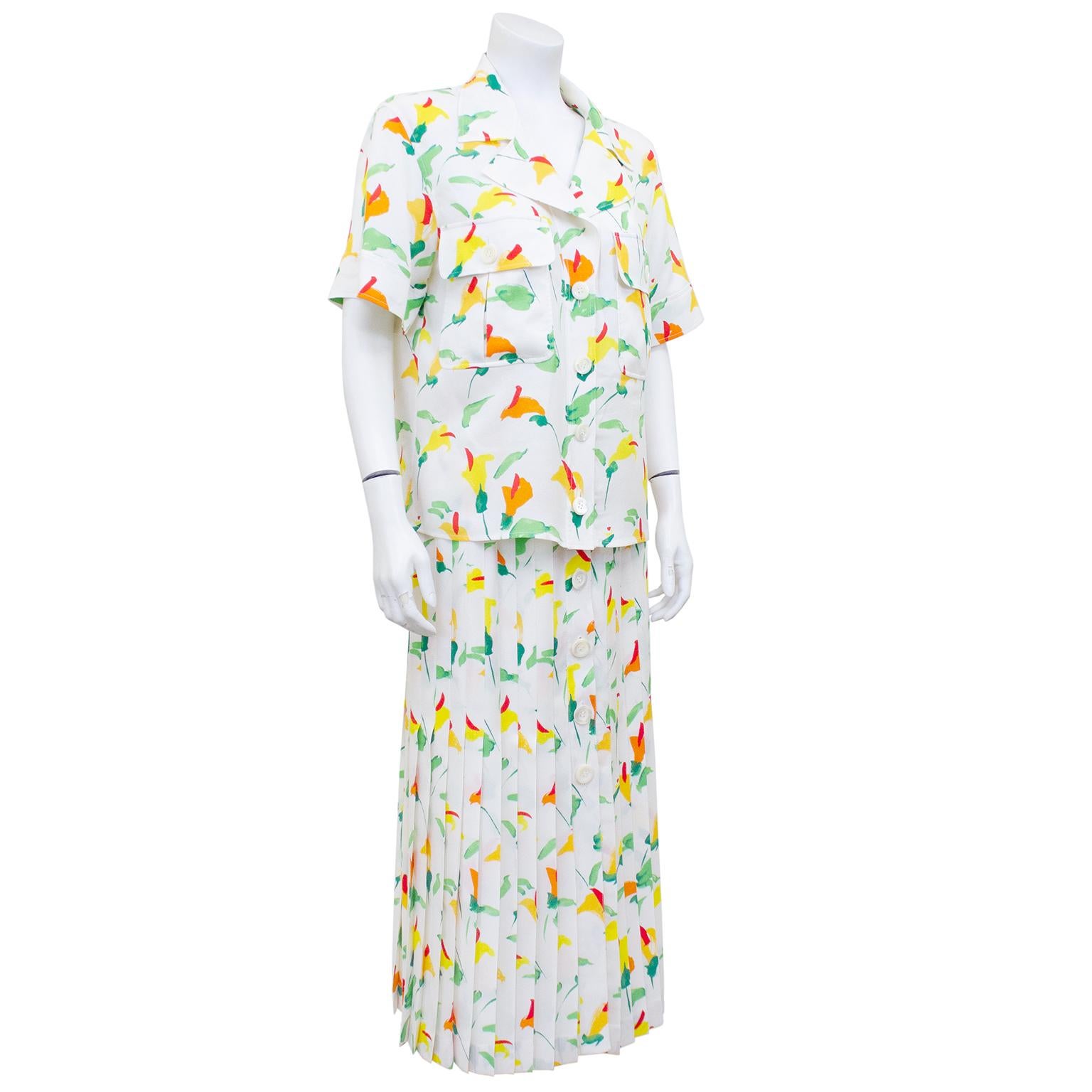 1980s white silk Valentino ensemble with allover bright calla lily print. Boxy short sleeve shirt with notched collar, large flap pockets and iridescent white plastic buttons. High waisted pleated midi skirt with matching button down centre front.