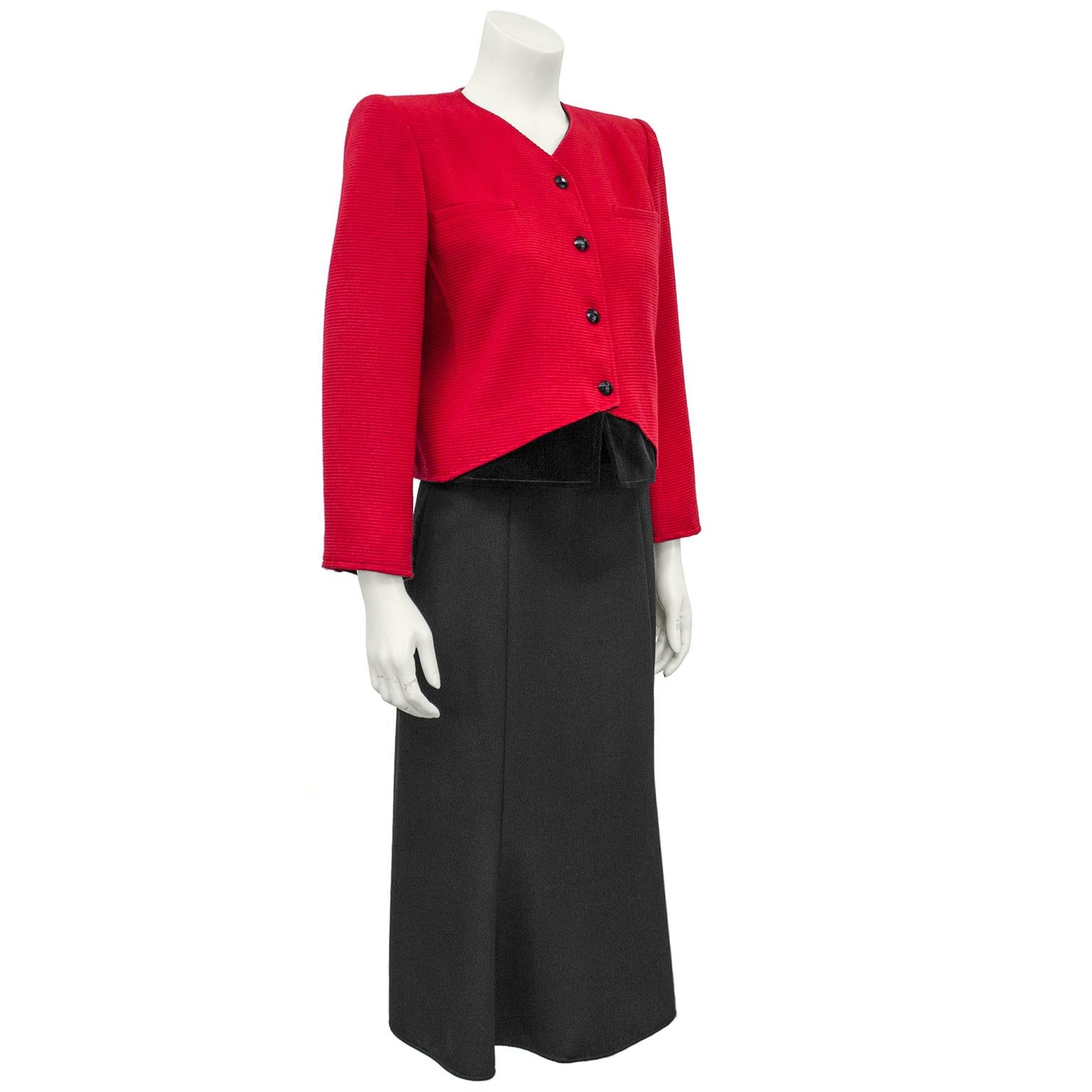 1980s Valentino midi skirt suit with velvet details. The collarless jacket is made of a horizontally ribbed red fabric with two front pockets on the bust and black faceted buttons down the front. The front hem of the jacket features a cut away