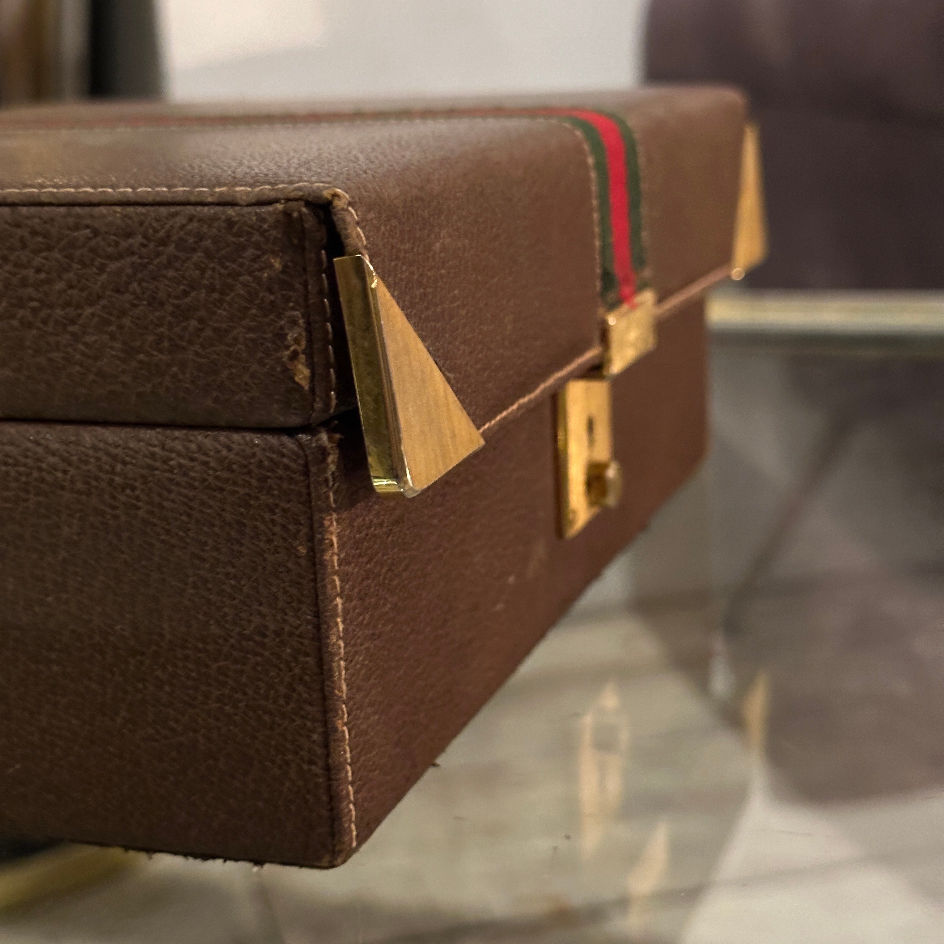 Se trata de un joyero vintage de cuero marrón de los años 80 de Gucci. Presenta un diseño rectangular estructurado con un exterior de cuero marrón intenso, acentuado por la icónica franja de red verde y roja de la marca que cruza la parte superior.