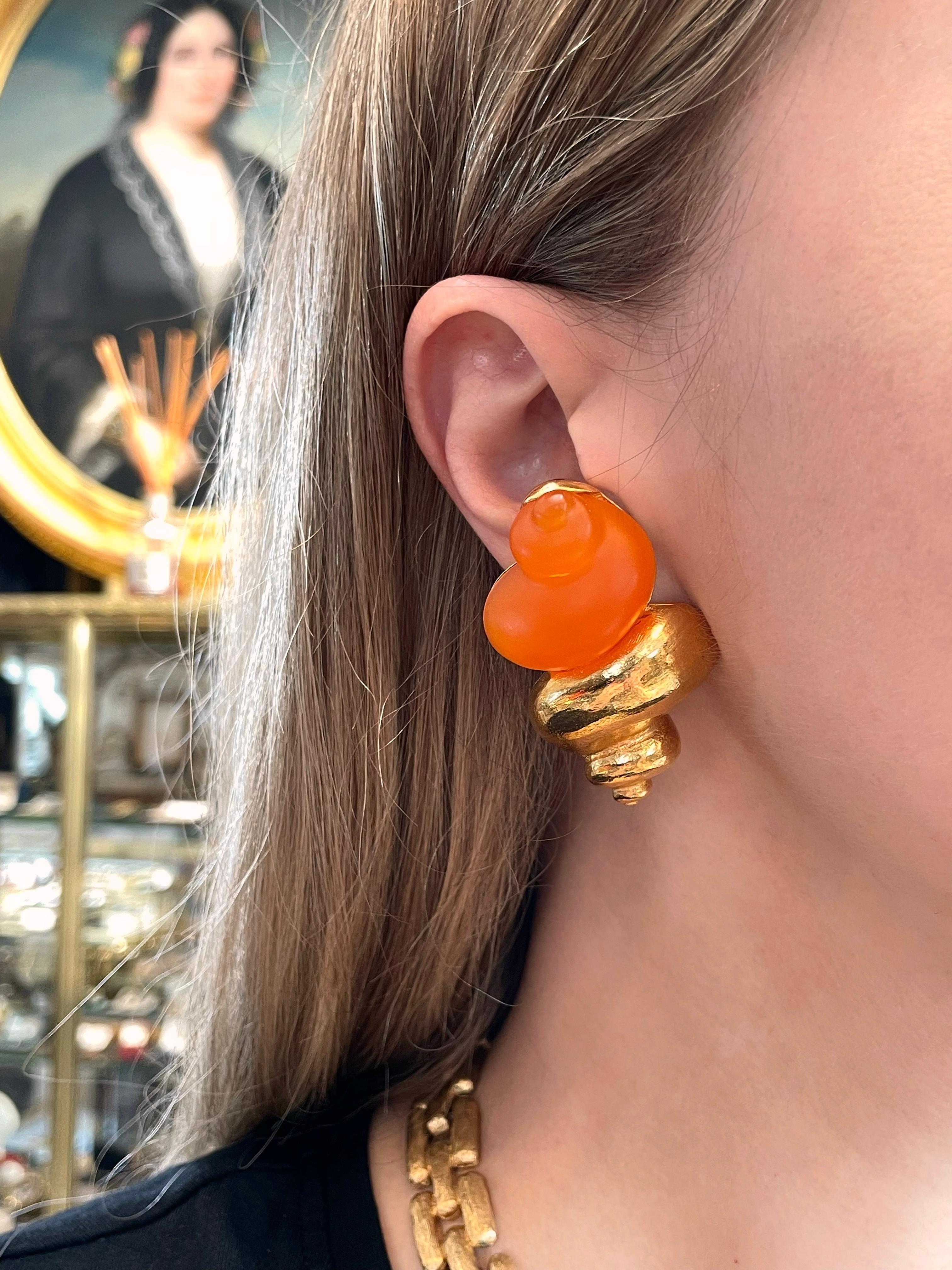 Si tratta di orecchini a clip a forma di conchiglia in tonalità oro disegnati da Christian Dior negli anni '80. Questo pezzo è placcato in oro e abbinato a lucite arancione. 

Marcature: 