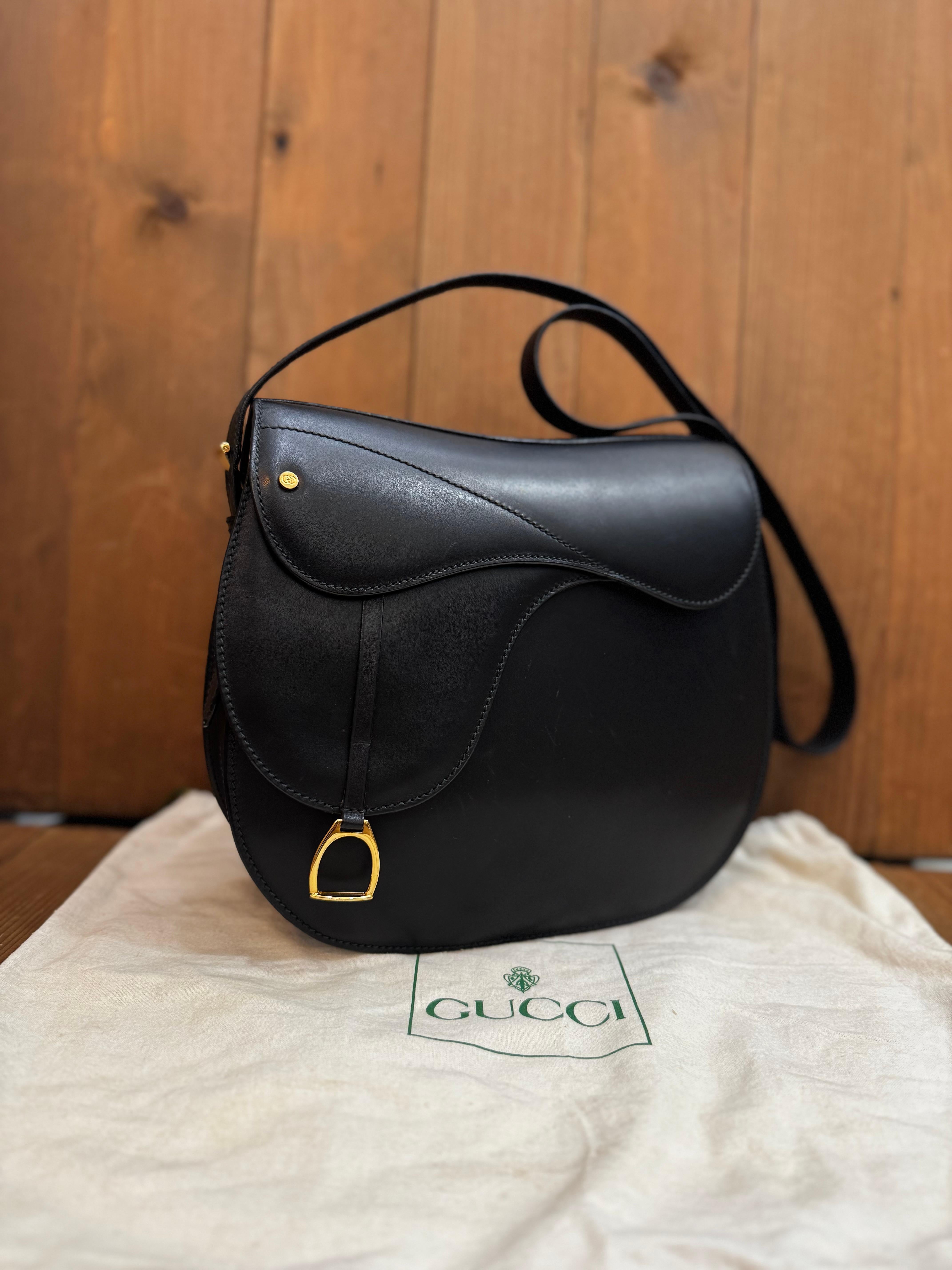 This vintage GUCCI saddle bag is crafted of smooth calfskin leather in black featuring gold toned hardware. Front magnetic snap closure decorated with a stirrup charm opens to a black leather interior featuring a zippered pocket. A rare quiet luxury