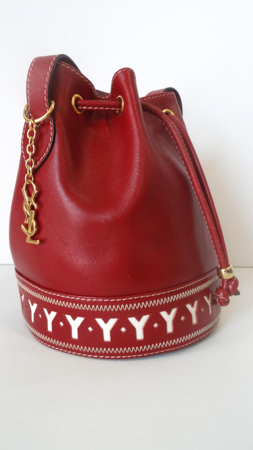 The Most Adorable Bag Is Here! Circa 1980s, this Yves Saint Laurent bucket bag is made of soft lipstick red pebble leather and features gold plated hardware. Includes contrasting white trim stitching and an adjustable belt style shoulder strap. The