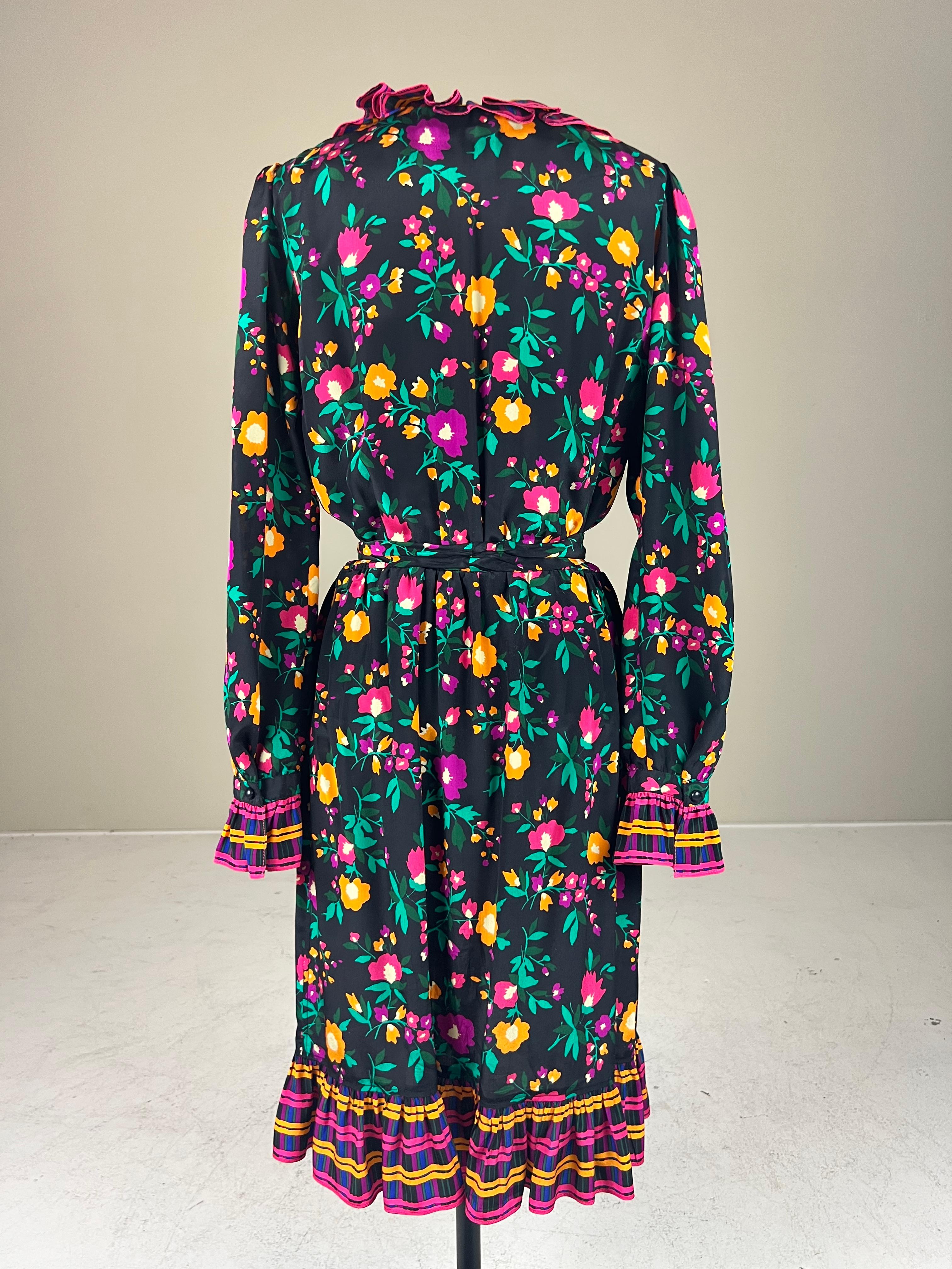 Cette robe cache-cœur en soie d'Yves Saint Laurent Rive Gauche de 1980* est dotée d'un magnifique imprimé floral multicolore. Les fleurs roses, orange et violettes entourées de feuilles vertes sont posées sur un fond noir. Au niveau de l'encolure,