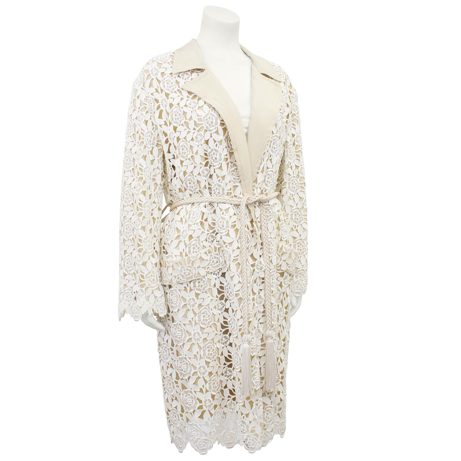 Stunning Yves Saint Laurent Rive Gauche coat from the 1980's. Beige silk with a cream floral guipoire lace overlay. Notched collar and scalloped edge at cuffs and hem. Faux horizontal slit pockets. Two belt loops with thin braided belt with tassel