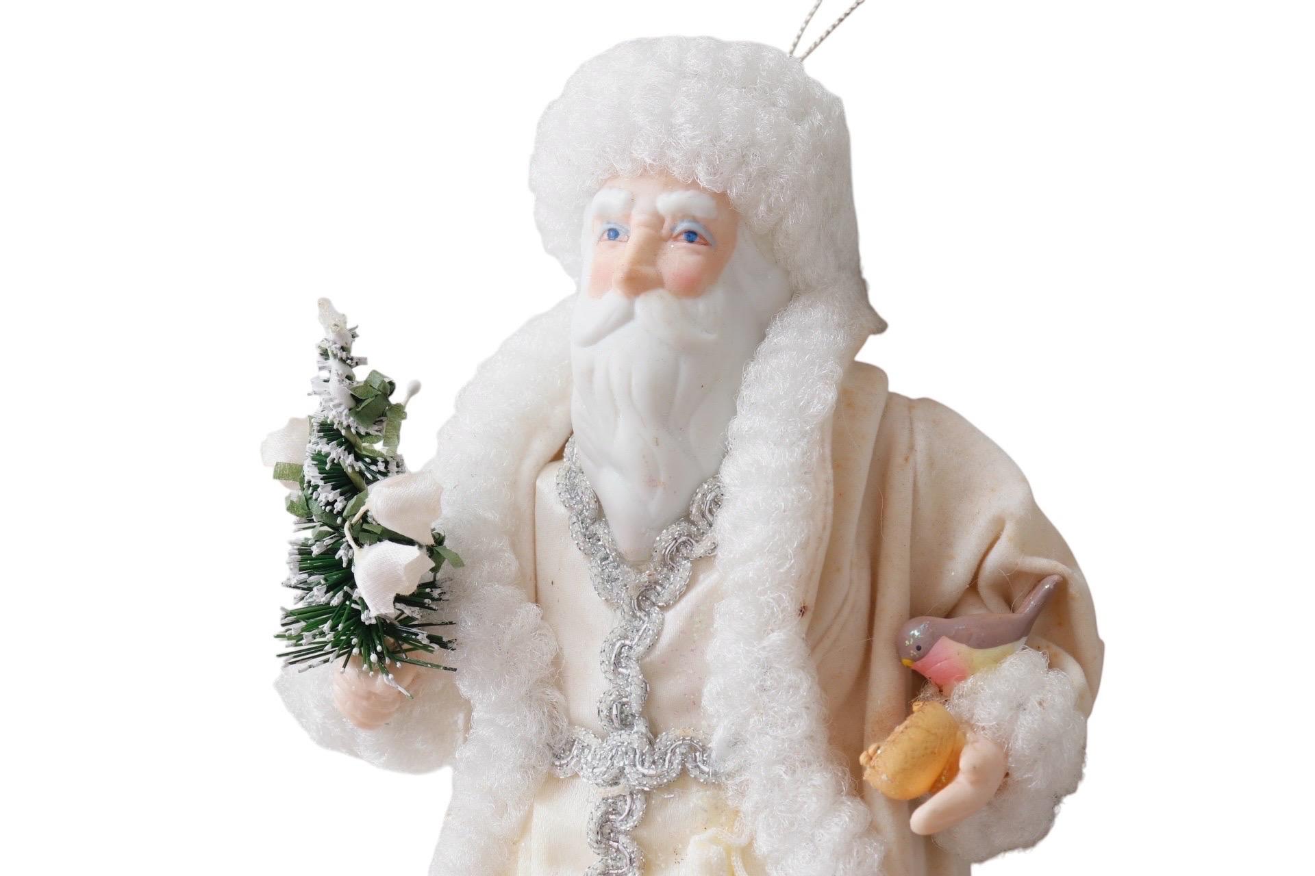 A porcelain Father Christmas hanging ornament by Kurt S. Adler for their Santa’s World collection. Santa wears a glittering white gown adorned in silver and a white fur trimmed robe. Porcelain legs are painted white to give the look of stockings and