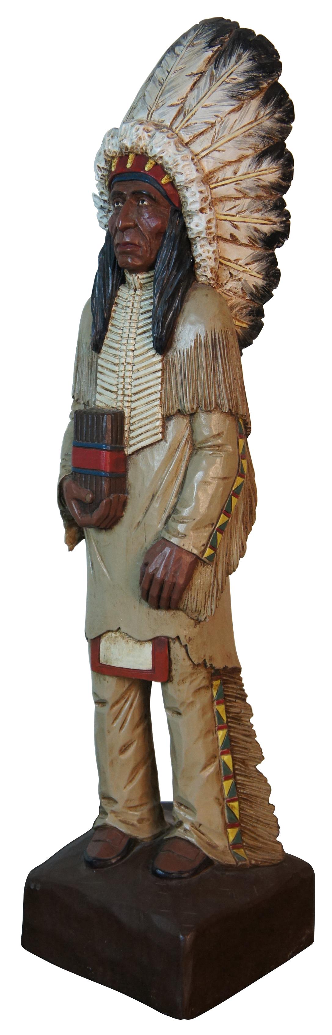 Vintage hand carved wood sculpture in the shape of a Native American man in a large feathered headdress, holding a package of cigars. Signed on the base “Sue Elliott 6/88.” The word “Weeks” is carved lightly on one side of the base. A fun mercantile