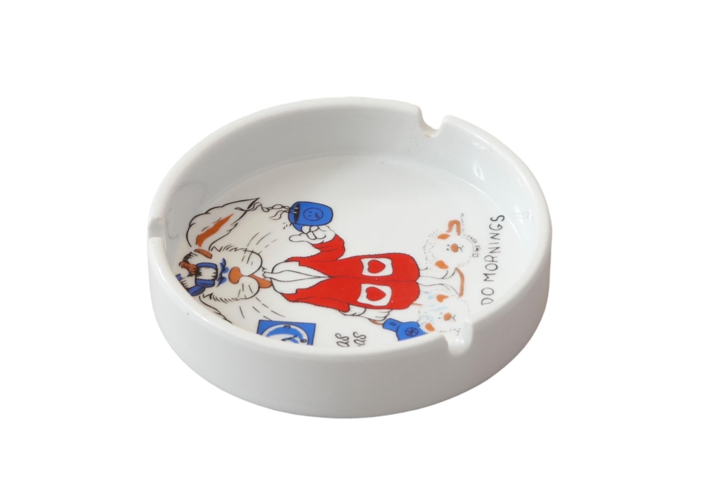 A round souvenir white ceramic ashtray, printed in the center with a rabbit in a dressing gown and slippers, hair dryer and coffee mug in hand, above the phrase “I don’t do mornings”. To the left is a clock showing 5:00 (presumably AM) above ‘Las