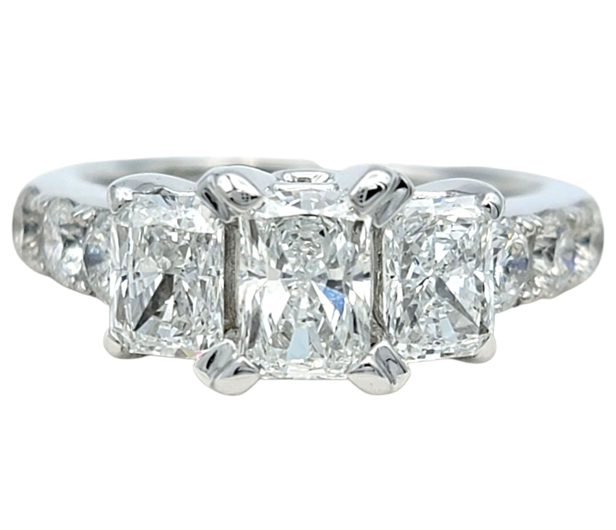 This elegant engagement ring showcases timeless brilliance with a radiant-cut diamond at the center, flanked by two matching radiant-cut diamonds on each side. The centerpiece gently tapers into a row of round brilliant diamonds that cascade halfway