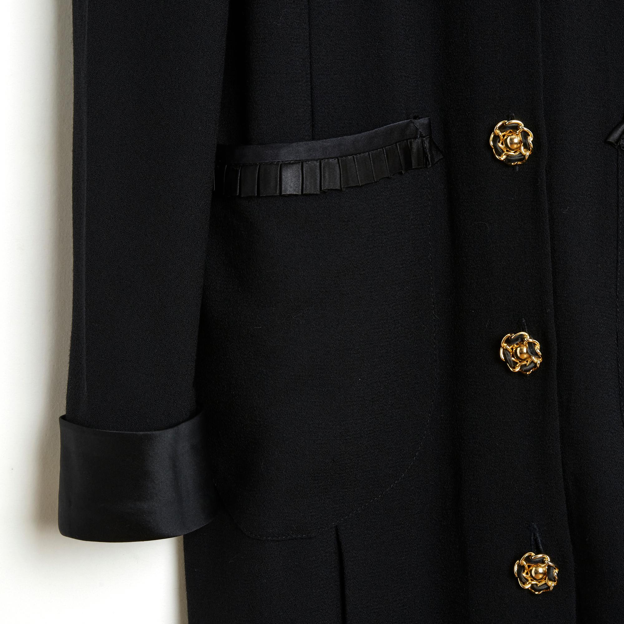 Chanel dress coat circa 1990 in black wool crepe, ankle length, high collar in wide black silk satin ribbon, closed in front with 7 buttons including 6 interwoven with black leather and 1 faceted highlighted with a brooch with the motif of a large