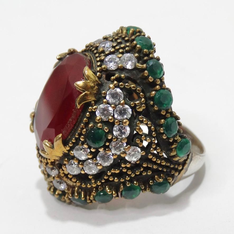 Stunning synthetic ruby and emerald cocktail ring circa 1990. This gorgeous jumbo ring features a synthetic ruby at the center surrounded by silver circular rhinestones and bronze and gold detailing. The ring is completed with an arrangement of