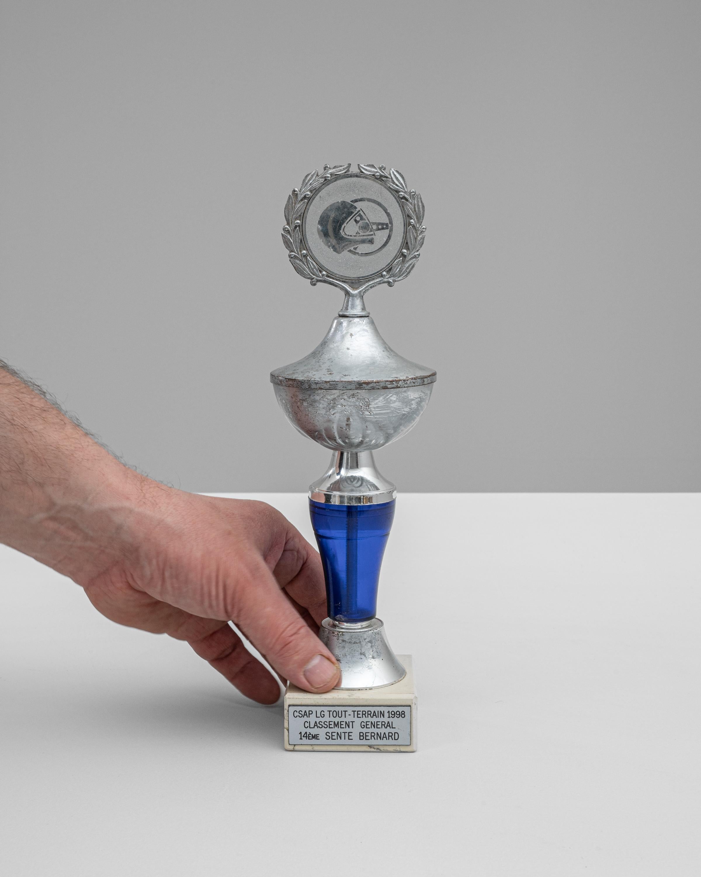 Dieser belgische Metall- und Marmorpokal aus den 1990er Jahren vom CSAP LG Tout-Terrain 1998 ist eine beeindruckende Auszeichnung, die die Kraft und Entschlossenheit des Geländesports repräsentiert. Das Design der Trophäe ist sowohl auffällig als