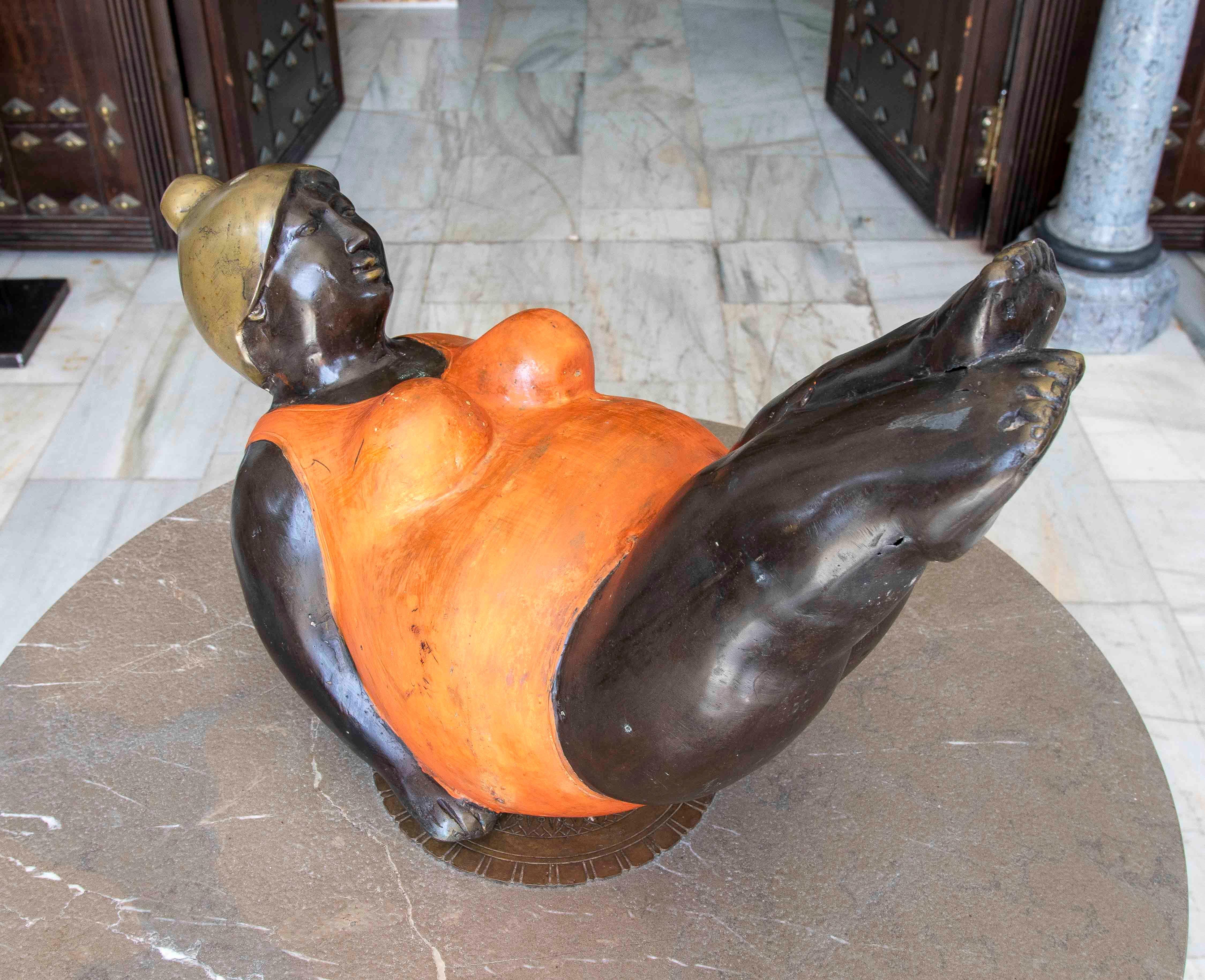 Contemporary patinated bronze sculpture depicting a generously sculpted woman reclining in a balanced position, her legs elevated. She wears a vibrant orange swimsuit and a gold bathing cap. This piece evokes an aesthetic close to that of sculptor