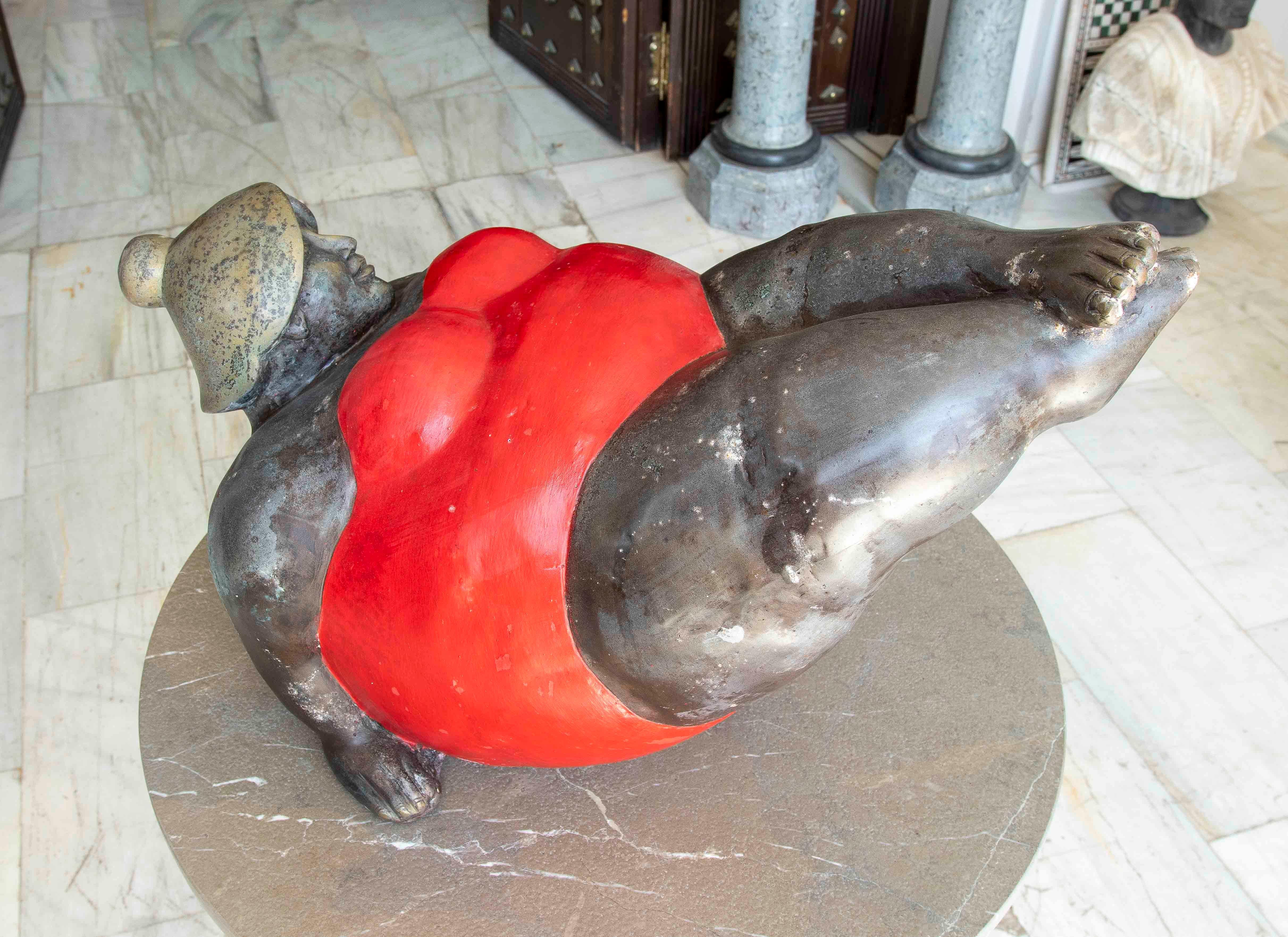Escultura contemporánea de gran expresividad que representa a una mujer de formas voluptuosas recostada, luciendo un llamativo traje de baño rojo. Elaborada en bronce patinado con detalles pintados a mano, esta pieza celebra la belleza en la