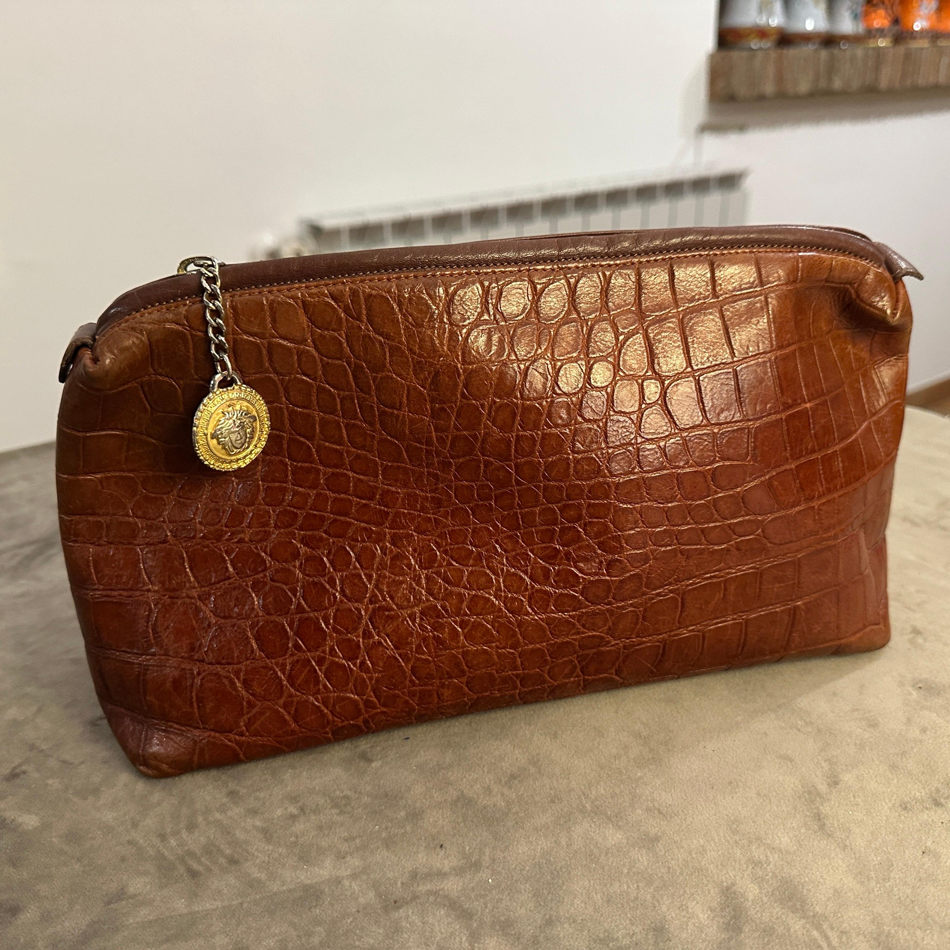 This is a vintage 1990s brown leather clutch bag by Gianni Versace. The bag is crafted from high-quality leather embossed with a crocodile pattern, giving it a luxurious and textured appearance. It features a rich, warm brown hue that enhances its