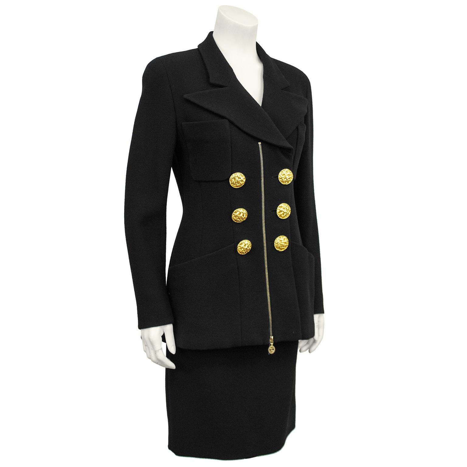 1990s Chanel black wool skirt suit. Double breasted jacket features an exaggerated collar, large woven gilt metal buttons, two small and two large patch pockets and a centre gold zipper. Zipper opens from bottom and can allow for a little more room