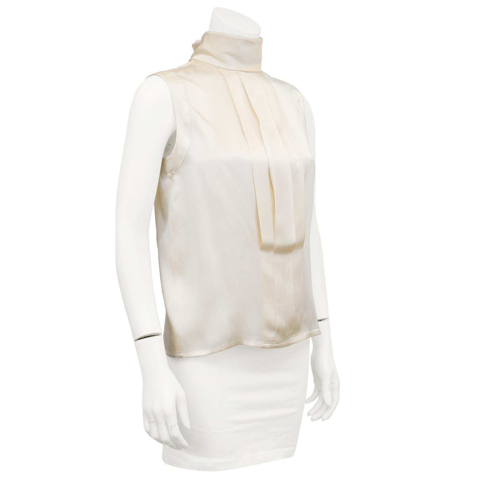 1990s Chanel cream silk blouse. Sleeveless with turtleneck collar. Flat pleated details at front. Button closures at back centre seam covered with pleat. French turned detail at nape of neck with small gold button engraved with the iconic Chanel