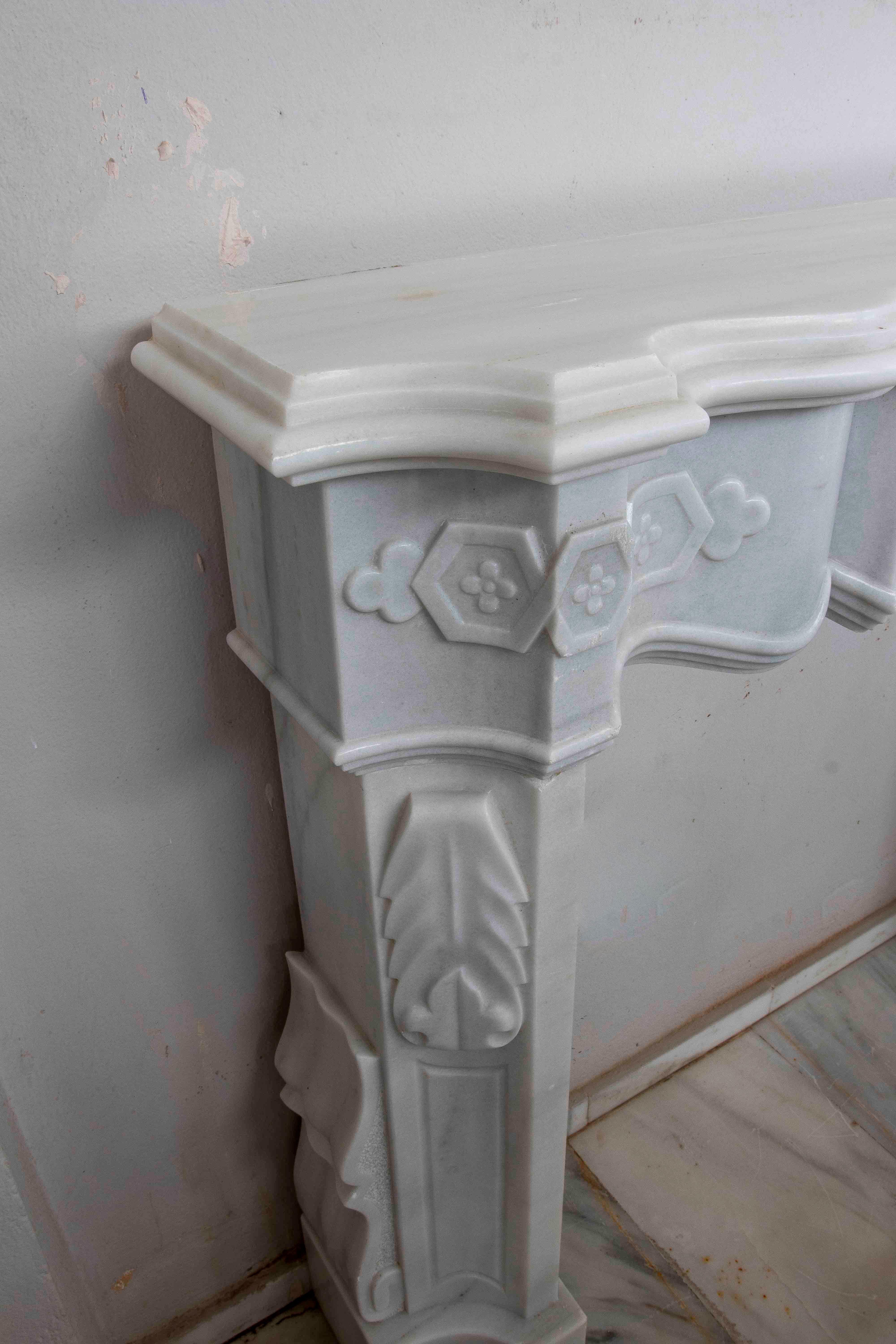 A sophisticated fireplace crafted from white marble with delicate, classically inspired ornamental carvings. Its design features a molded upper mantel with wavy lines, while the front displays floral motifs, acanthus leaves, and geometric elements