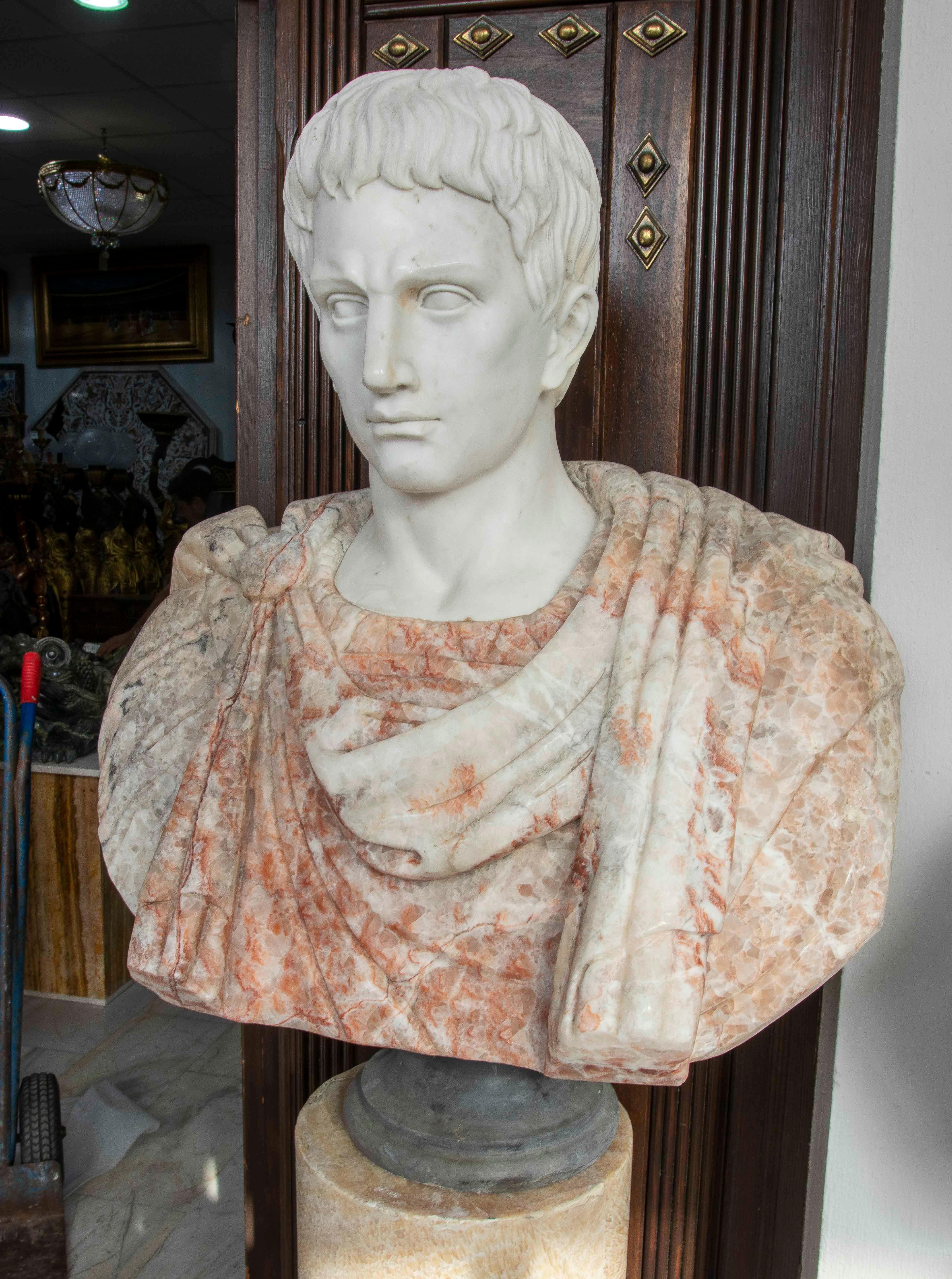 Impressionante busto in marmo che raffigura una figura romana dal portamento solenne. Il volto, realizzato in puro marmo bianco, contrasta con il busto rivestito in marmo con sfumature rosse e arancioni, creando un effetto visivo elegante e
