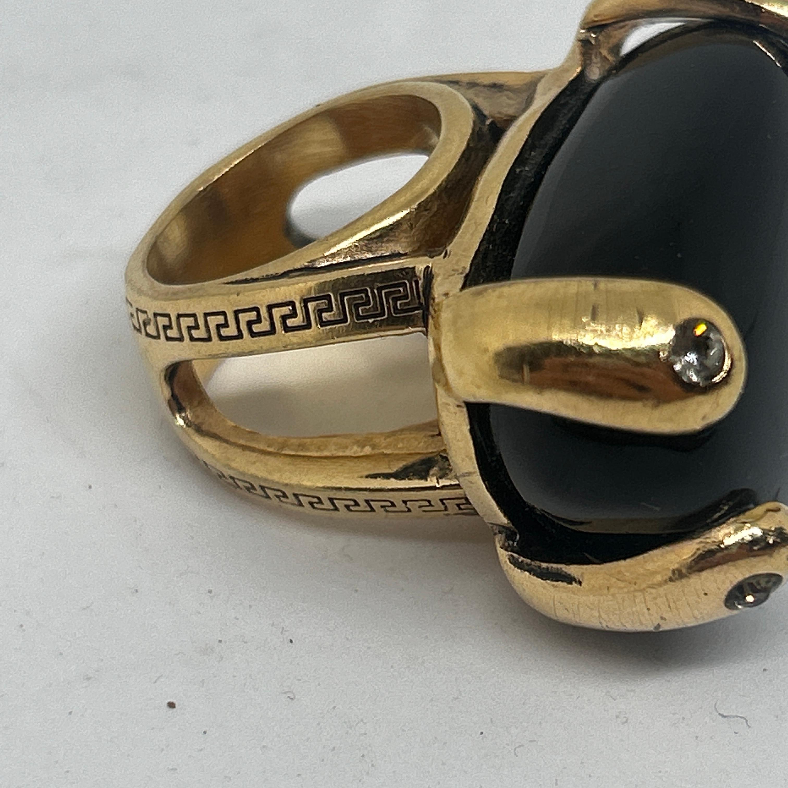 Dieser Gianni Versace-Cocktailring besticht durch sein kühnes Design mit einem großen, ovalen schwarzen Achat (als Herzstück). Der schwarze Achat ist glatt und poliert und bildet einen auffälligen Kontrast zu der **goldfarbenen Metallfassung**, die