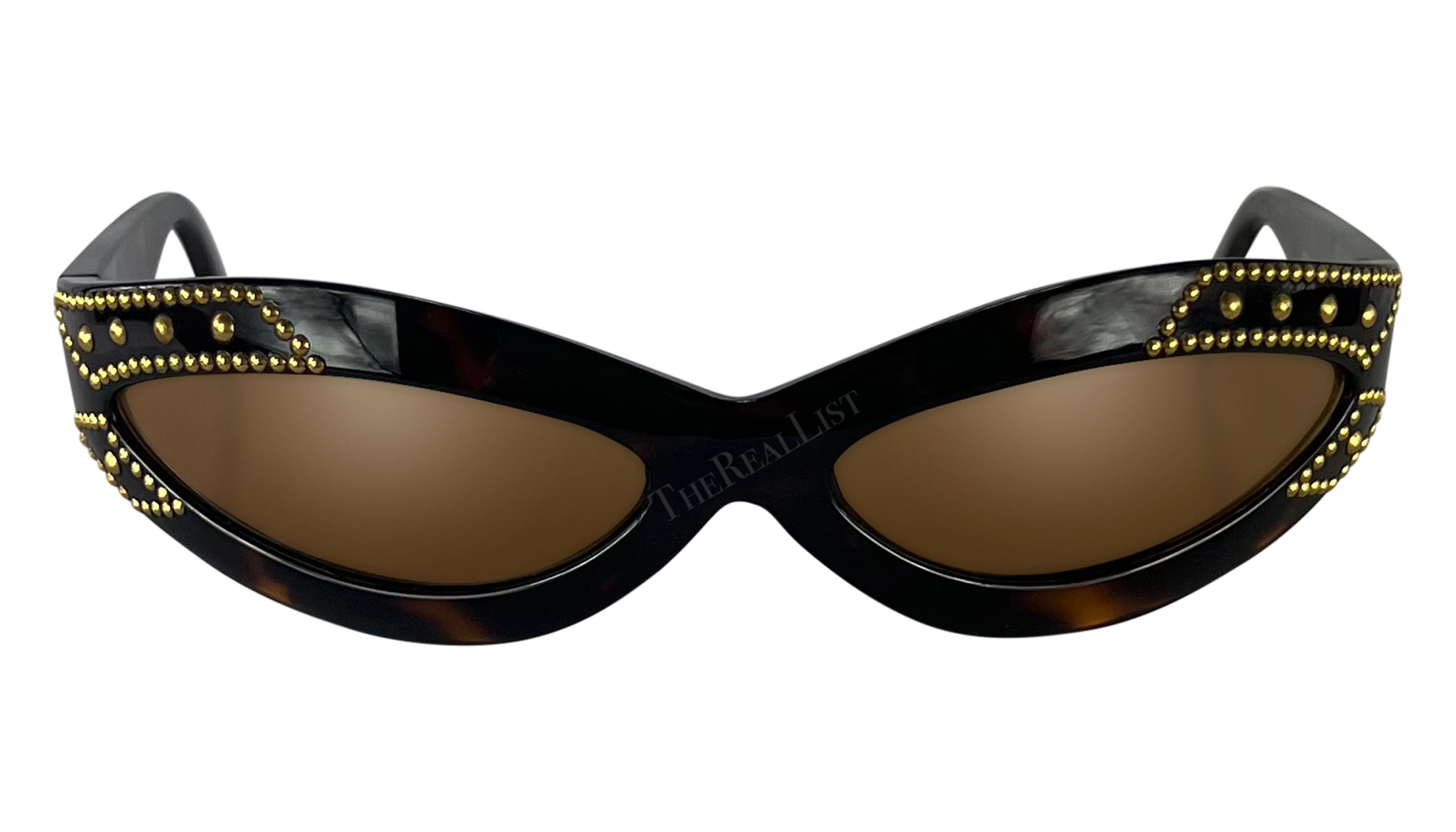 Datant des années 1990, ces lunettes de soleil ovales en fausse écaille, conçues par Gianni Versace, présentent un style enveloppant accentué par des clous dorés et un logo Versace Medusa sur un côté. 

Mesures approximatives-
Hauteur du cadre :