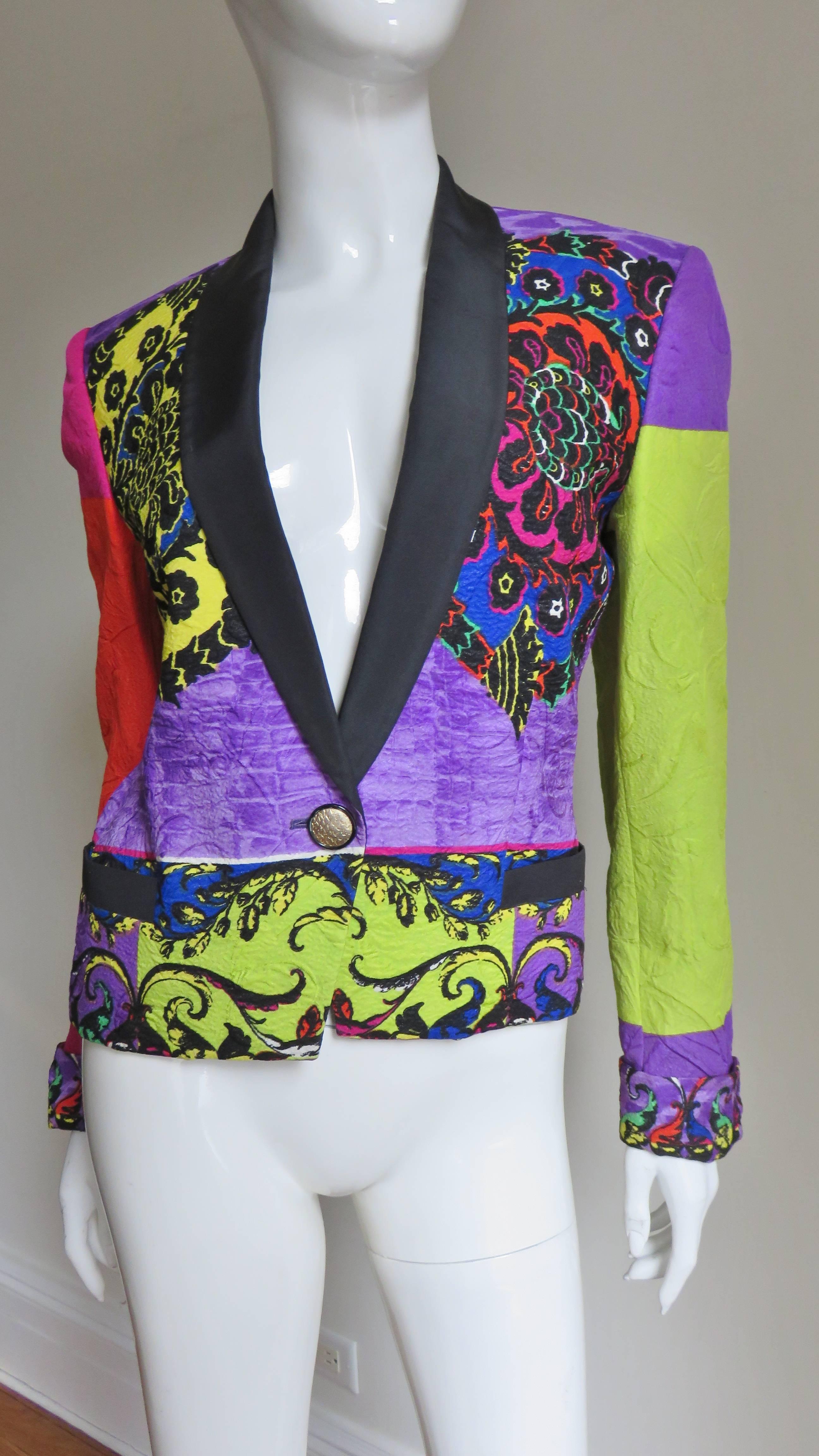 A fabulous brightly colored silk jacket from Gianni Versace in orange, lime, purple, magenta and black in elaborate patterns of flowers and abstract shapes.  It has a black silk shawl collar matching pockets at the hips.  It closes with one silver