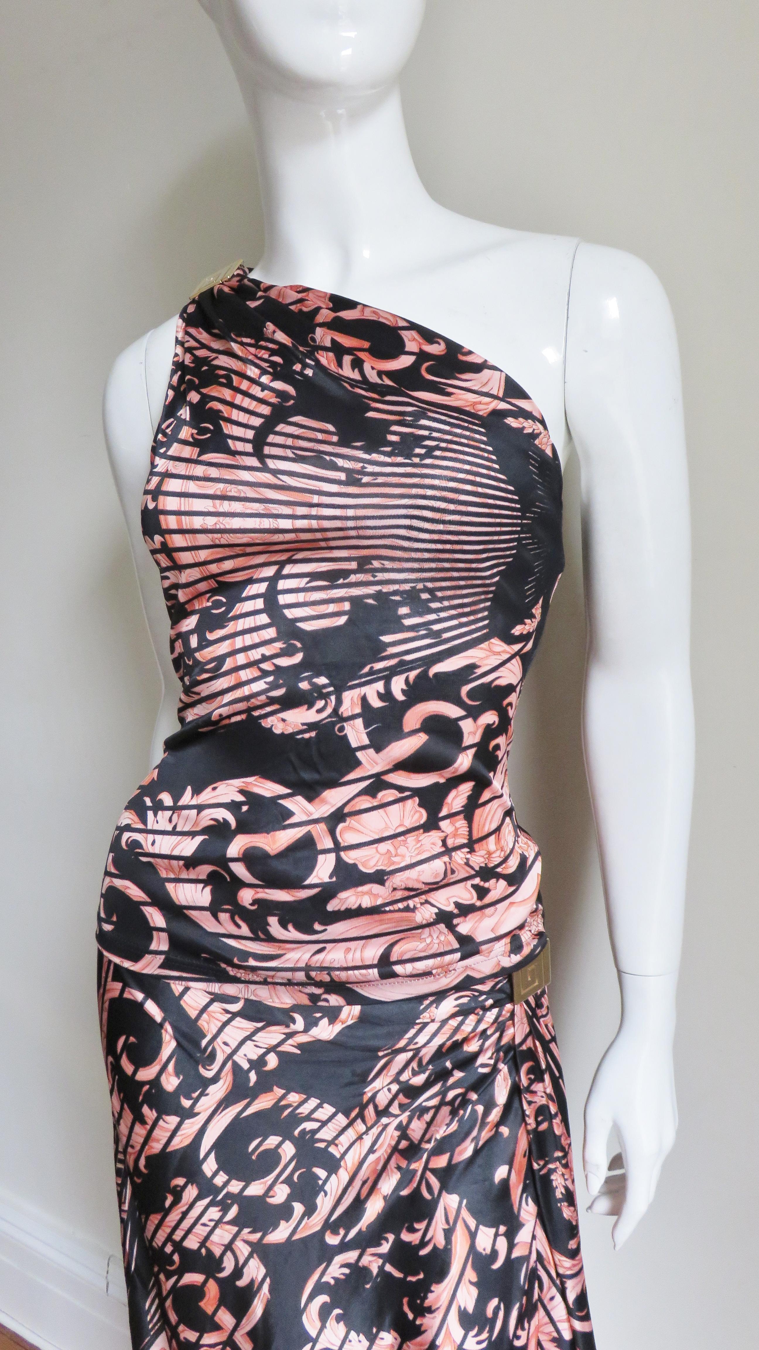 A fine silk jersey one shoulder top and matching skirt from Gianni Versace Couture in pink scrolls and lines print on a black background.  The top is fitted and has a gold metal signature Versace Greek key hardware at the one shoulder, the full