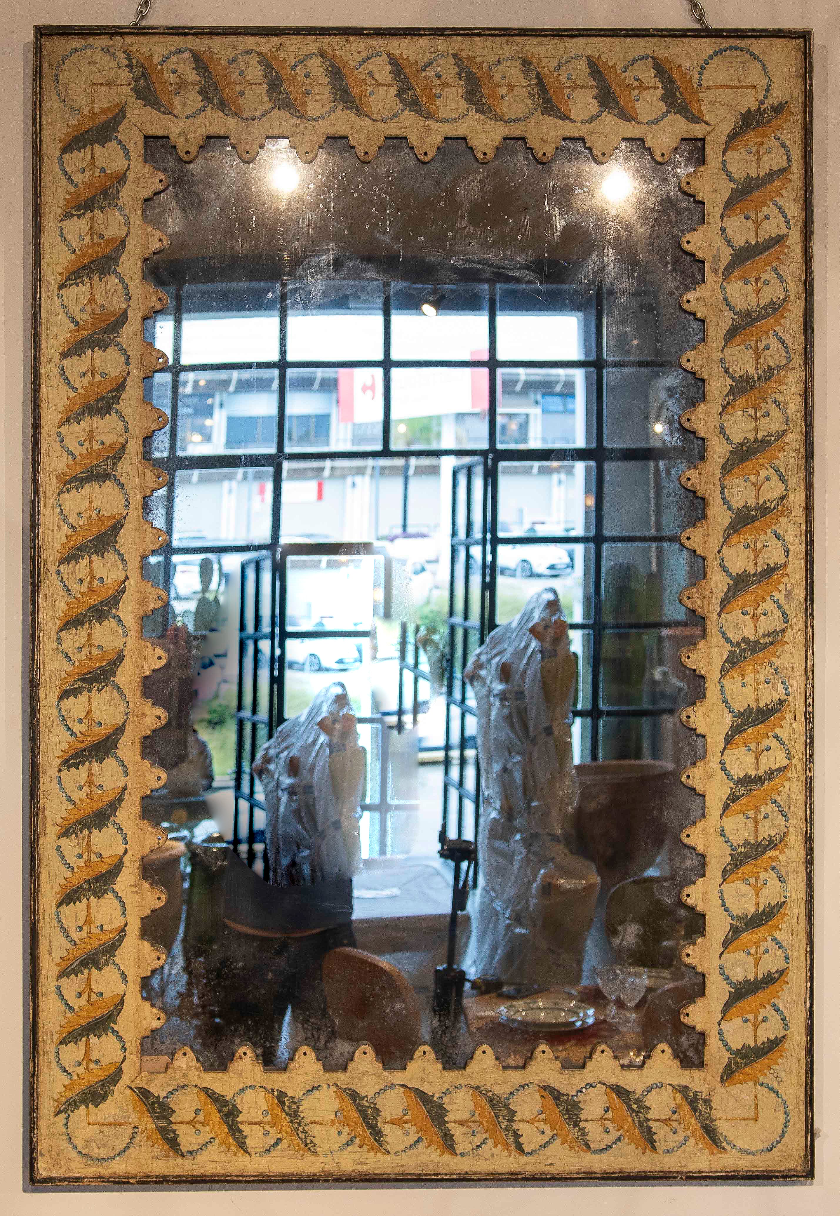 Elegante specchio in stile antico con cornice in legno intagliata e decorata a mano, caratterizzata da un disegno ornamentale con motivi vegetali e geometrici nei toni dell'oro, del blu e dell'ocra. La sua finitura invecchiata e la patina naturale