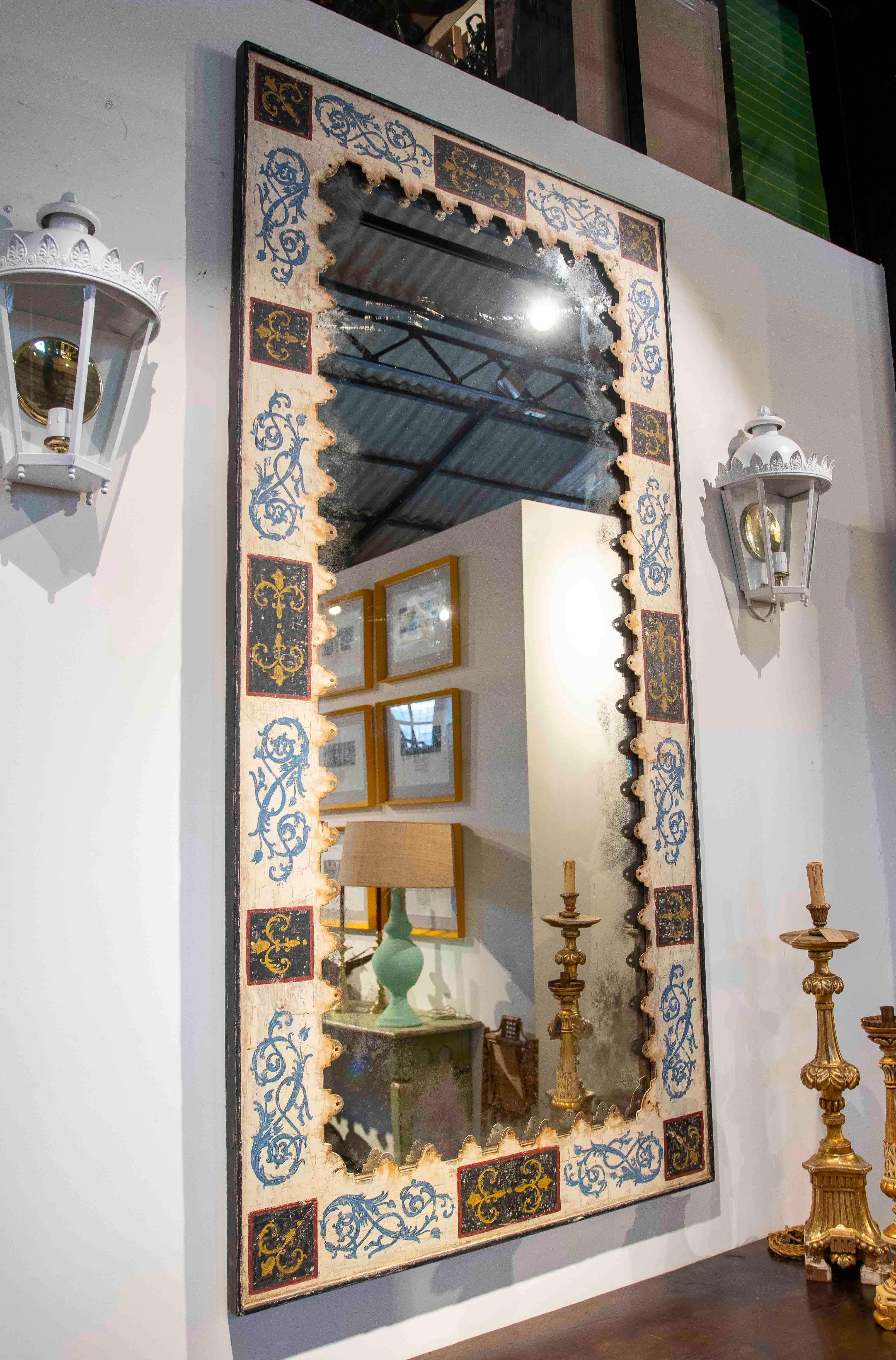 Elegante espejo con un marco de metal decorado con detalles pintados a mano en tonos azules, negros y dorados. Su diseño muestra un fino trabajo artesanal con motivos florales y arabescos, realzando su estética clásica y sofisticada. La pátina