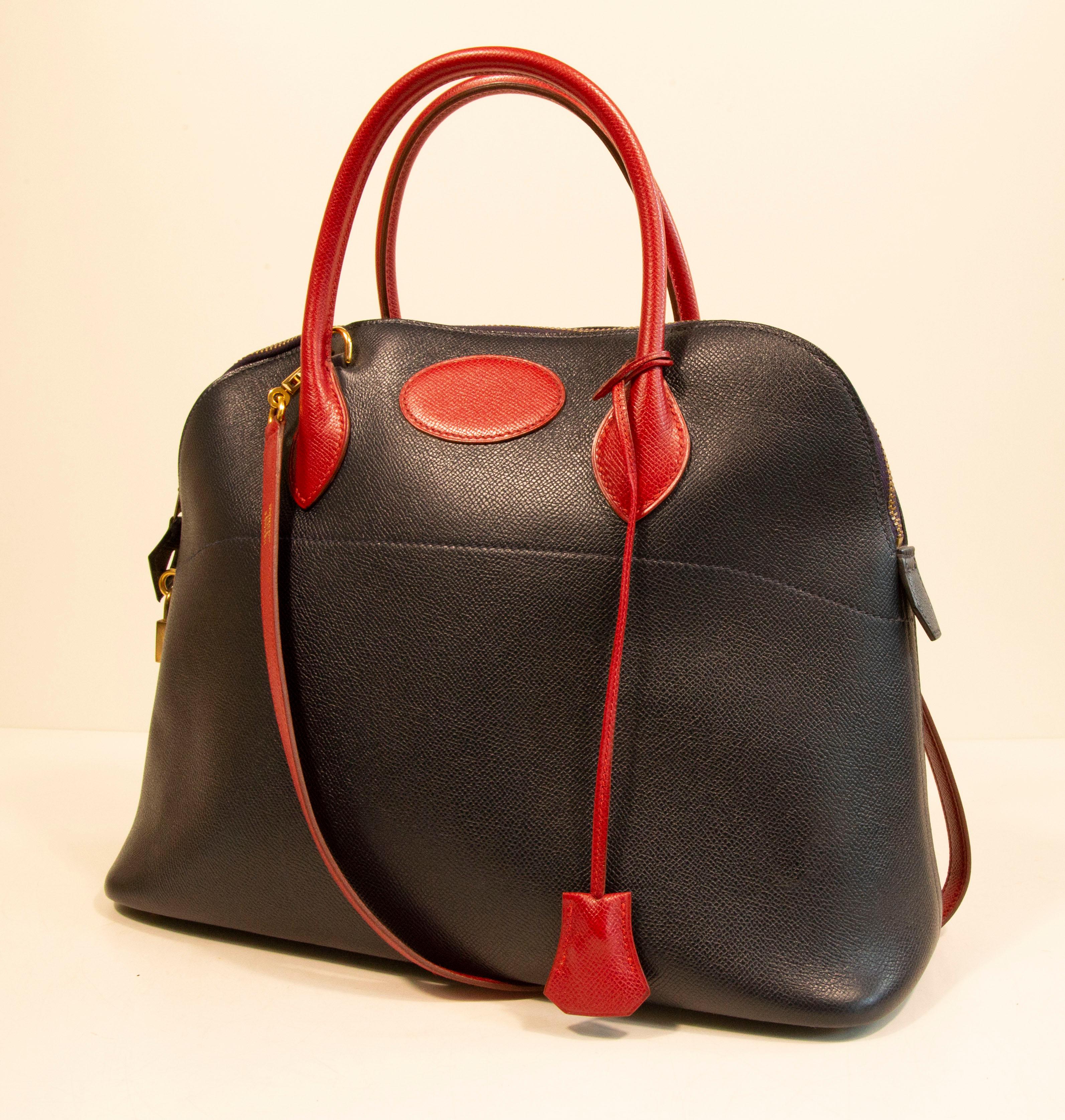 Hermes Bolide 35 handbag made of textured navy blue leather with red leather finish. The hardware is gold toned. The interior is lined with smooth navy blue leather. The interior consists of one major compartment   with one slide pocket. 
The bag