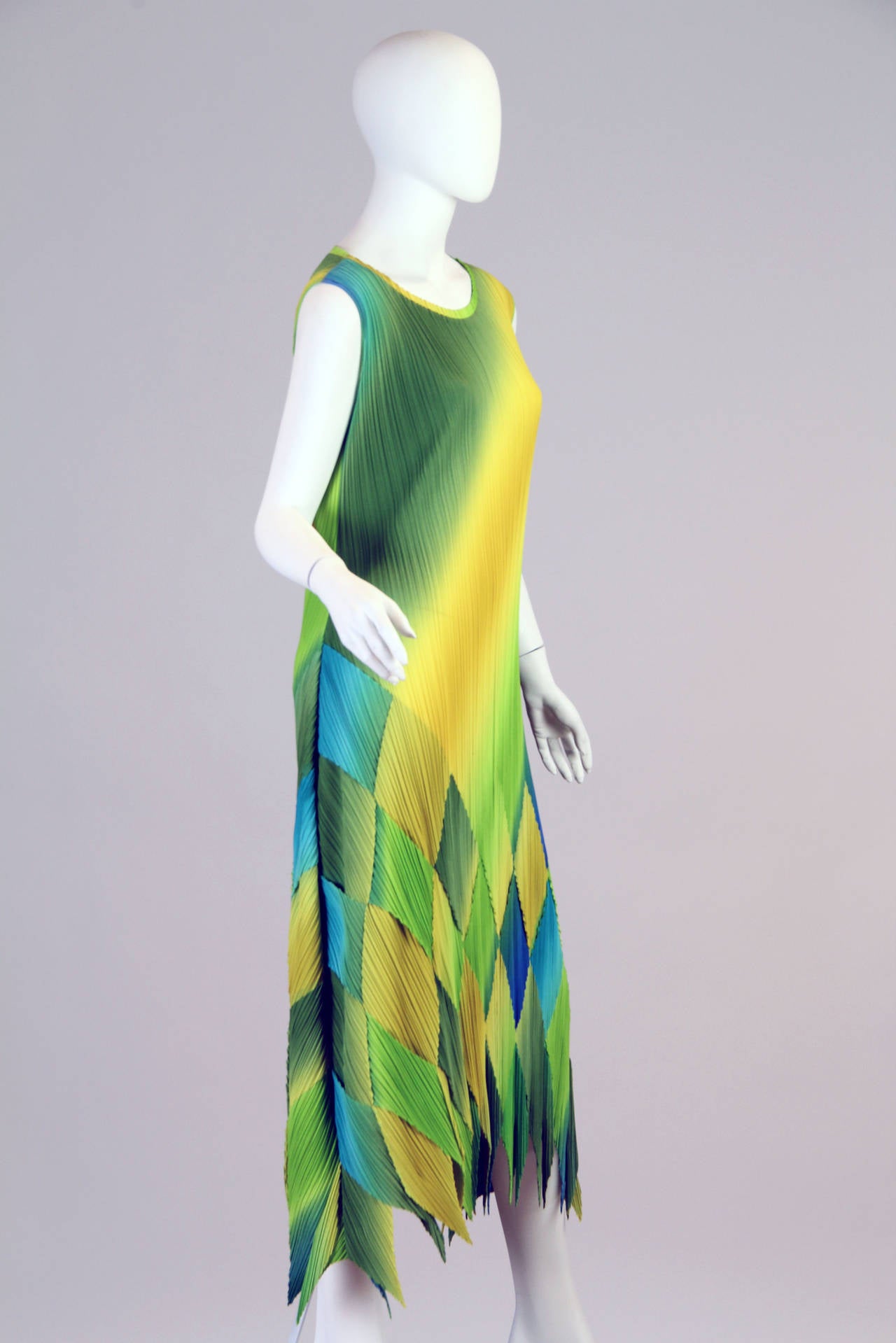 This 1990s Issey Miyake sleeveless dress is constructed of a vibrant a blue, green, and yellow pleated polyester fabric. The bright hues are enveloped together in a diamond pattern at the bottom of the dress, while a jagged hemline adds a chic flair