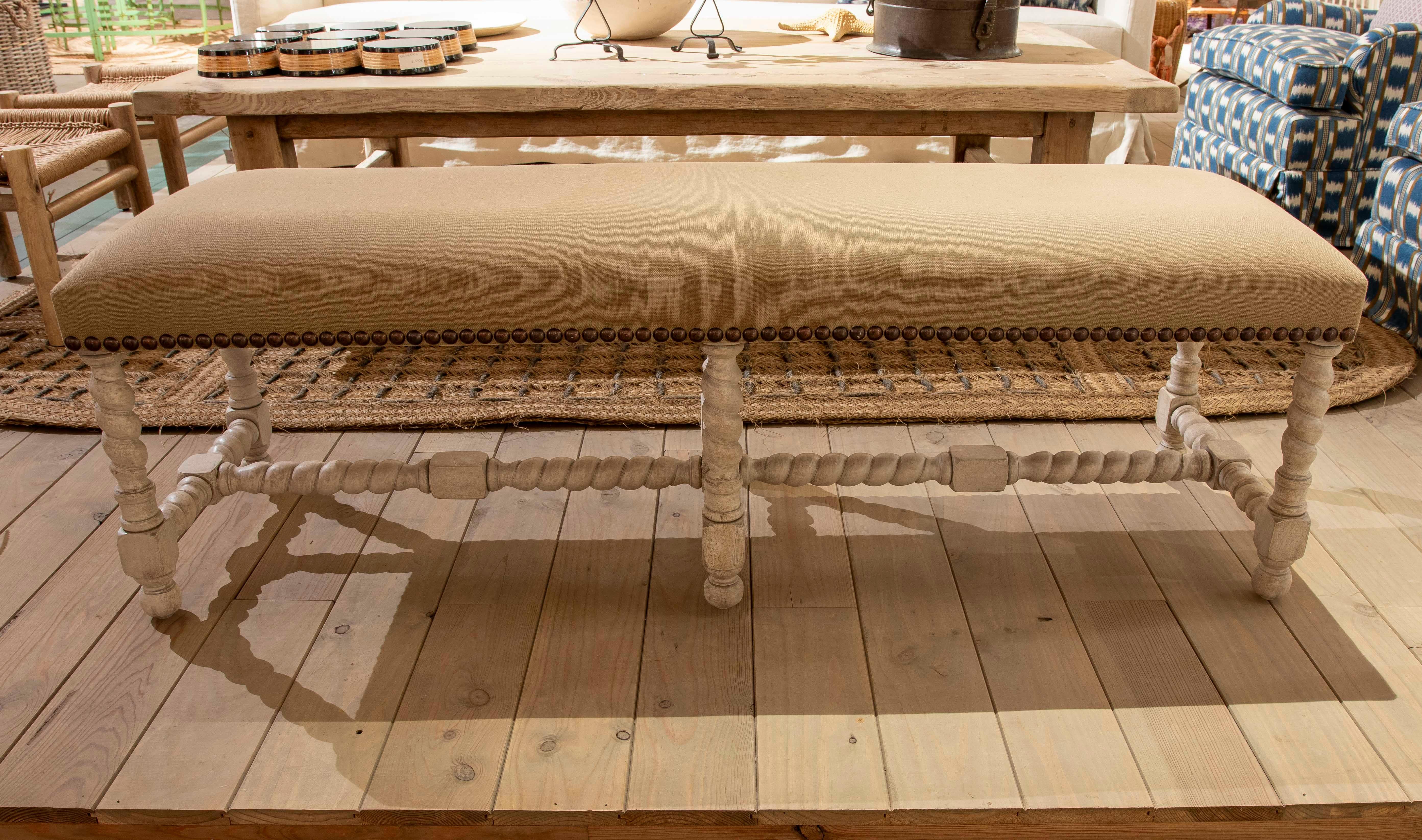 Banc sophistiqué de style classique avec une assise allongée tapissée de tissu beige et rehaussée de clous décoratifs le long du bord inférieur. La base est fabriquée en bois sculpté avec une finition décapée blanche, avec des pieds tournés et des