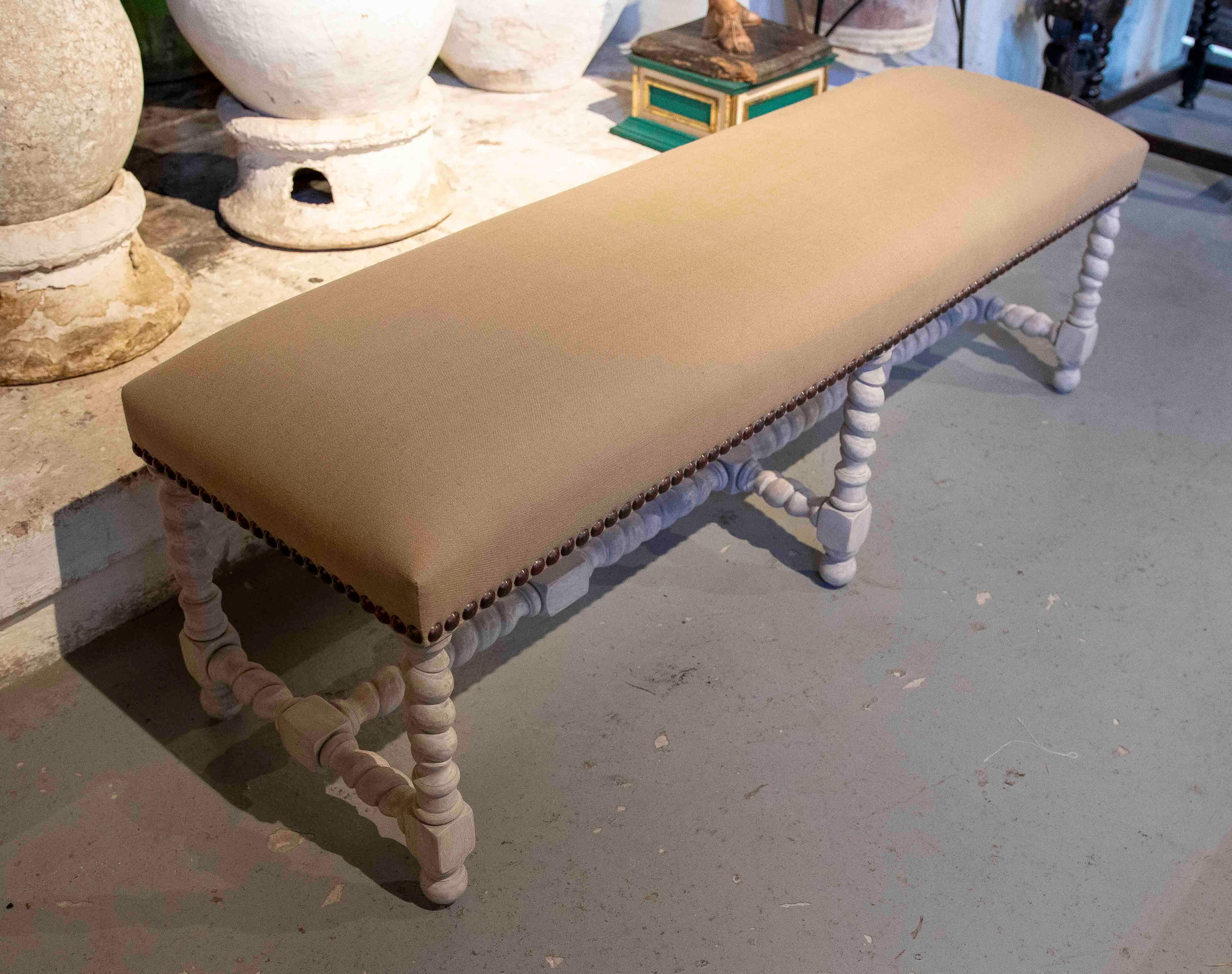 Elegant banc surdimensionné avec une structure en bois et des pieds tournés et croisés, finis dans un ton antiquaire clair. L'assise est recouverte d'un tissu taupe uni, terminé par une rangée de clous métalliques décoratifs. Une pièce polyvalente