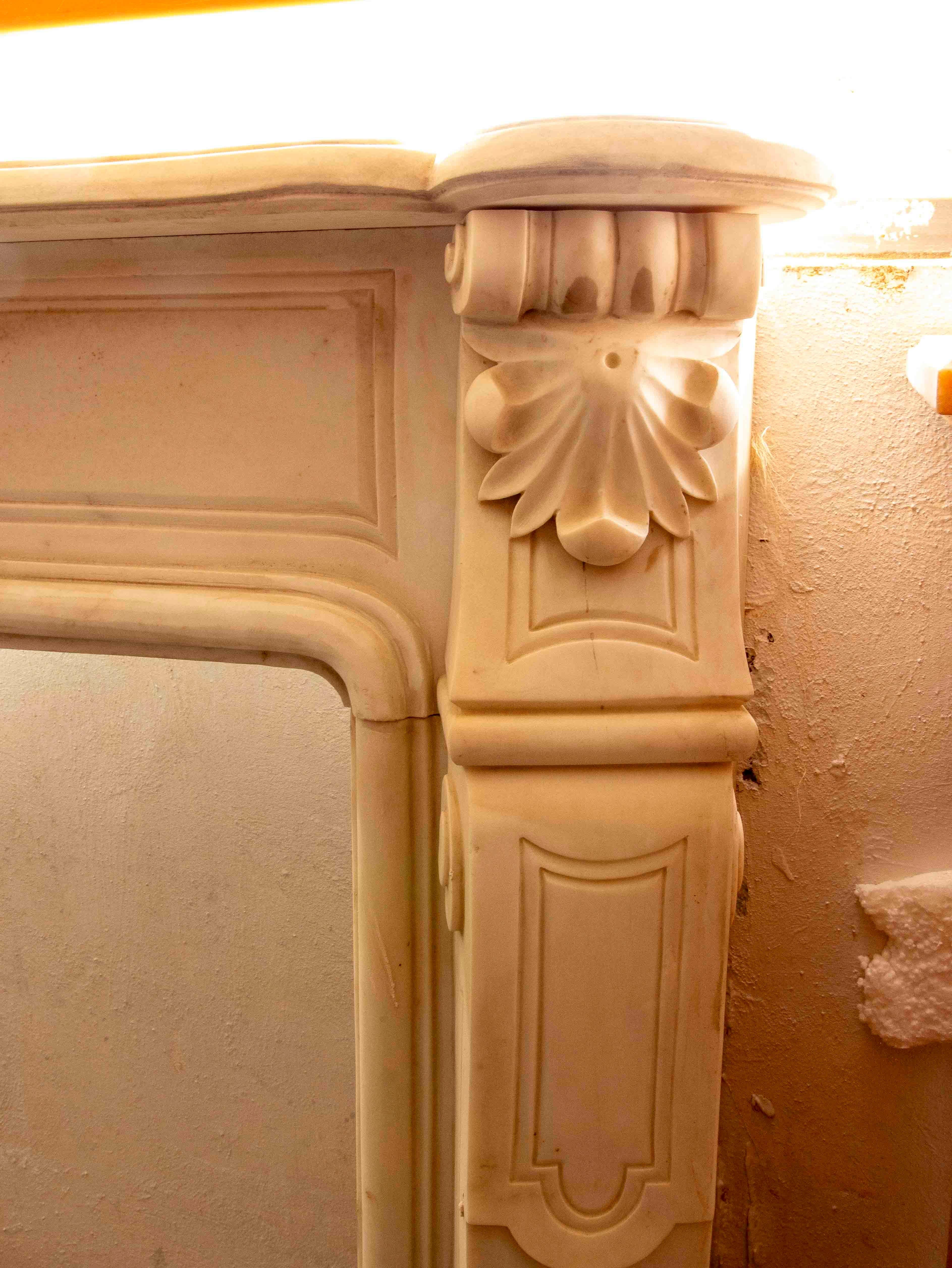 A sumptuous, hand-carved white marble fireplace in the Louis XV style, featuring flowing lines and delicate reliefs that evoke the elegance of the French Rococo. Its design includes a central medallion surrounded by floral motifs and scrolls,