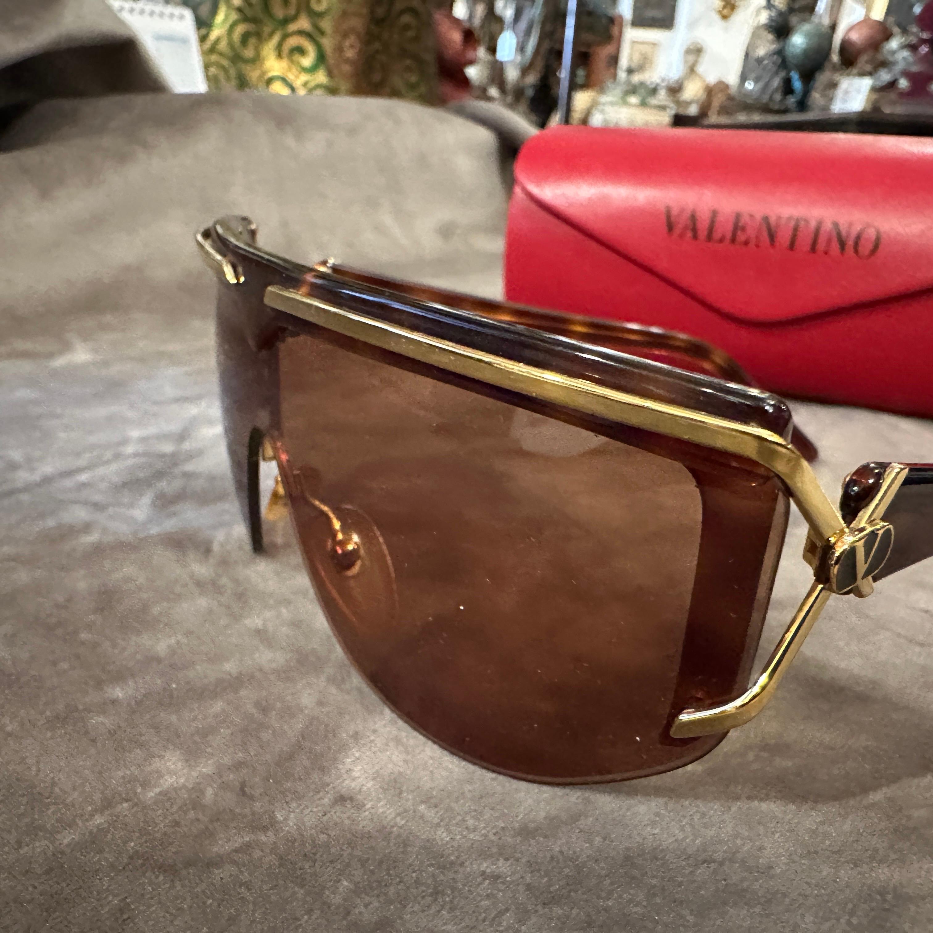 These vintage luxury oversized mask sunglasses by Valentino, dating back to the 1990s, exude bold sophistication and futuristic elegance. The design features a single, sweeping brown-tinted lens that seamlessly spans the entire front, creating a