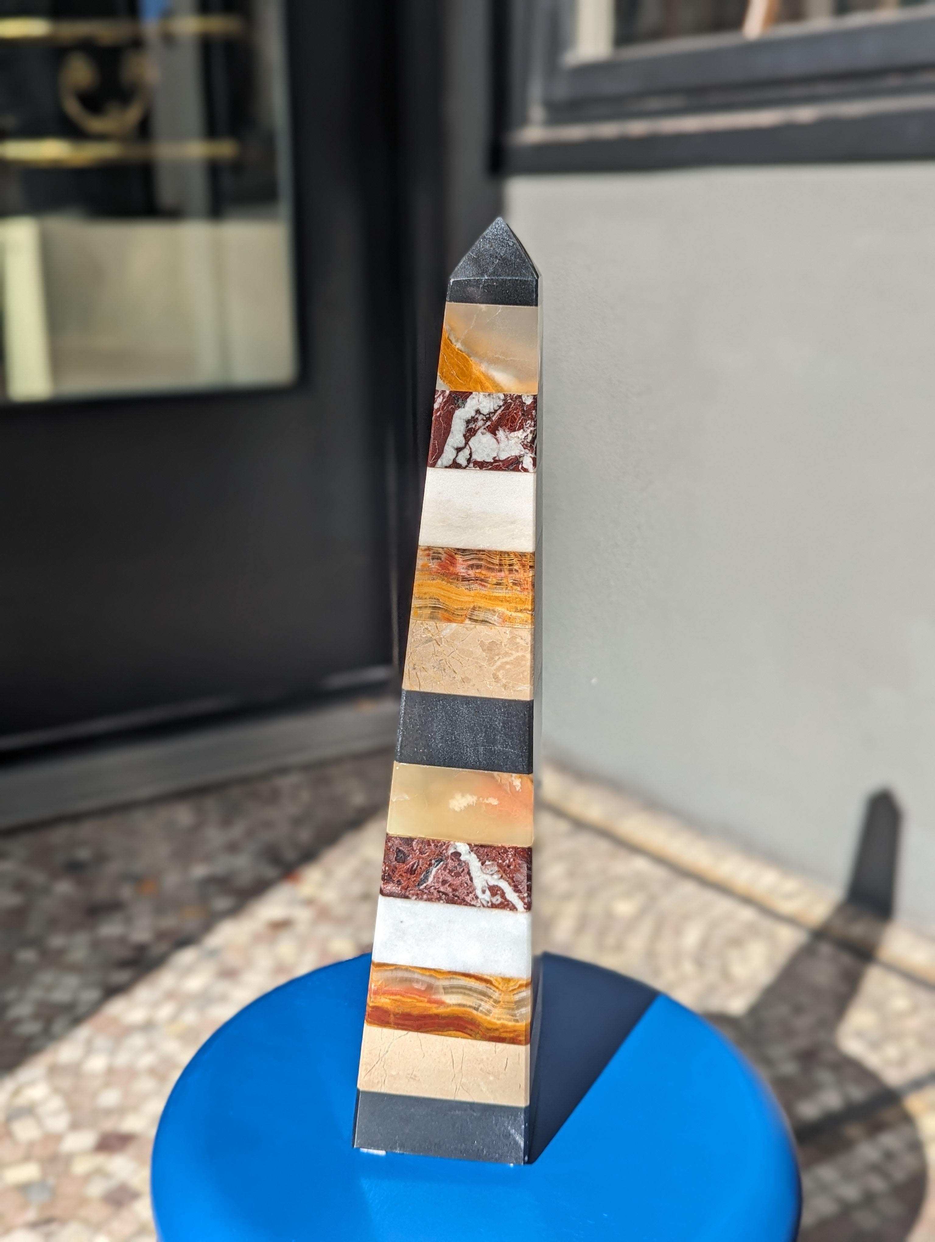 Step into a world where sleek lines meet natural splendor with this 1990s Modern Mixed Stone Geometric Obelisk. Imagine its vibrant onyx, travertine, and marble layers catching the light, casting playful shadows across your bookshelf. This isn't