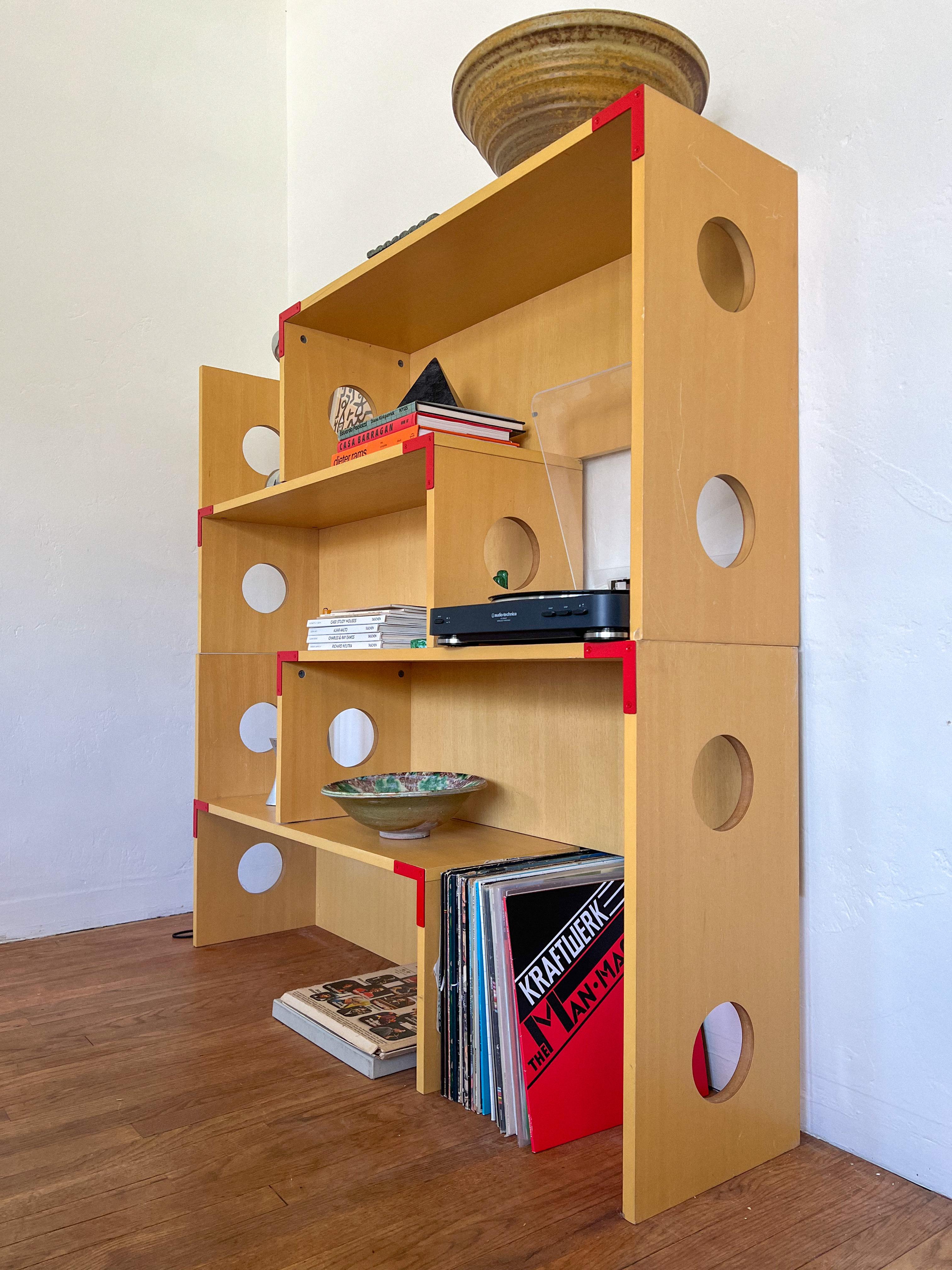 Una llamativa estantería modular al estilo de Philippe Starck o del diseño posmoderno de principios de los 90, con un acabado en laminado de abedul natural con atrevidos detalles rojos en las esquinas y juguetones recortes circulares por todas