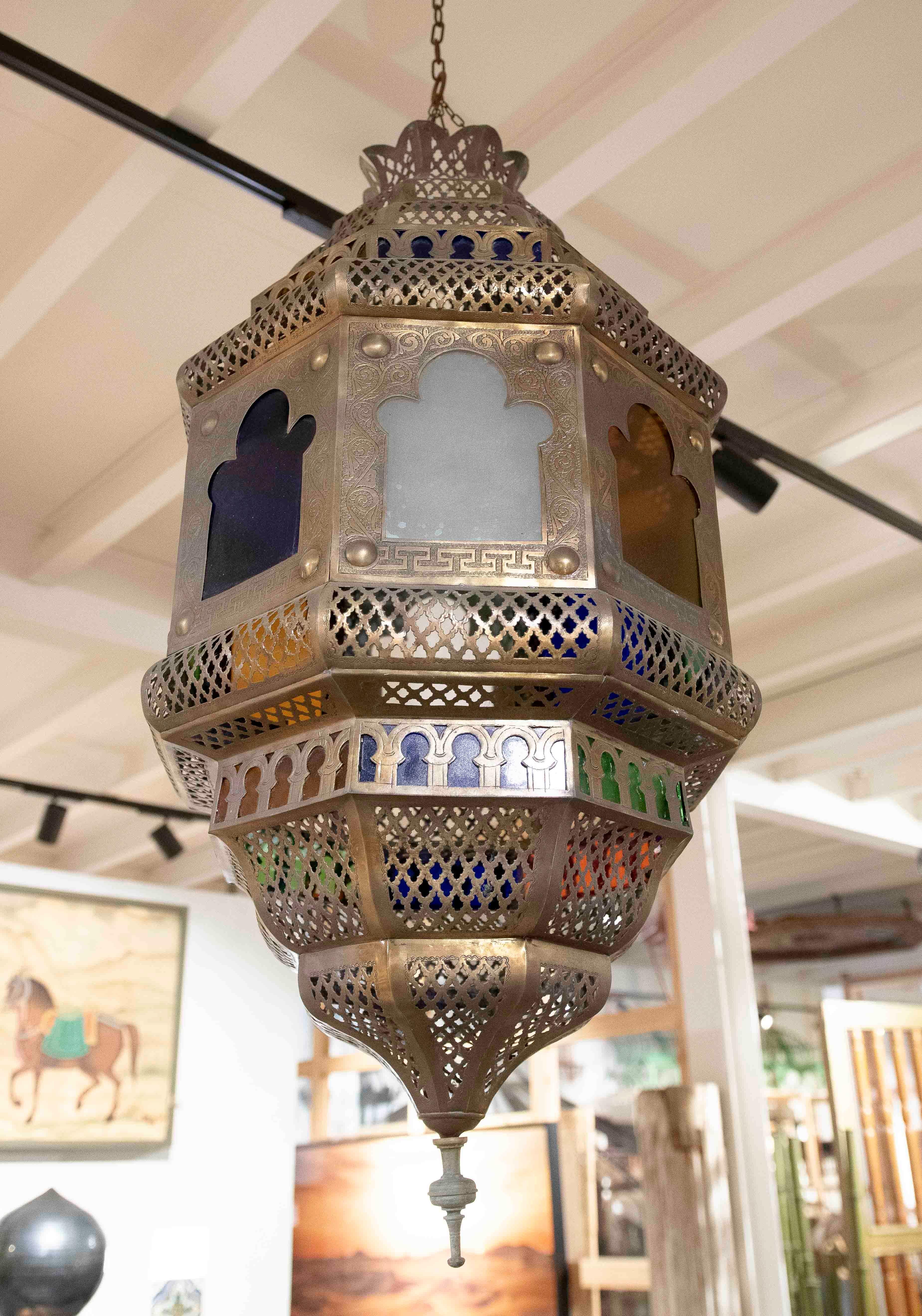 Stupenda lanterna da soffitto a sospensione dal design marocchino, realizzata in ottone traforato con intricati dettagli decorativi e pannelli di vetro in colori vivaci come verde, blu, ambra e bianco. La sua forma ottagonale e la finitura