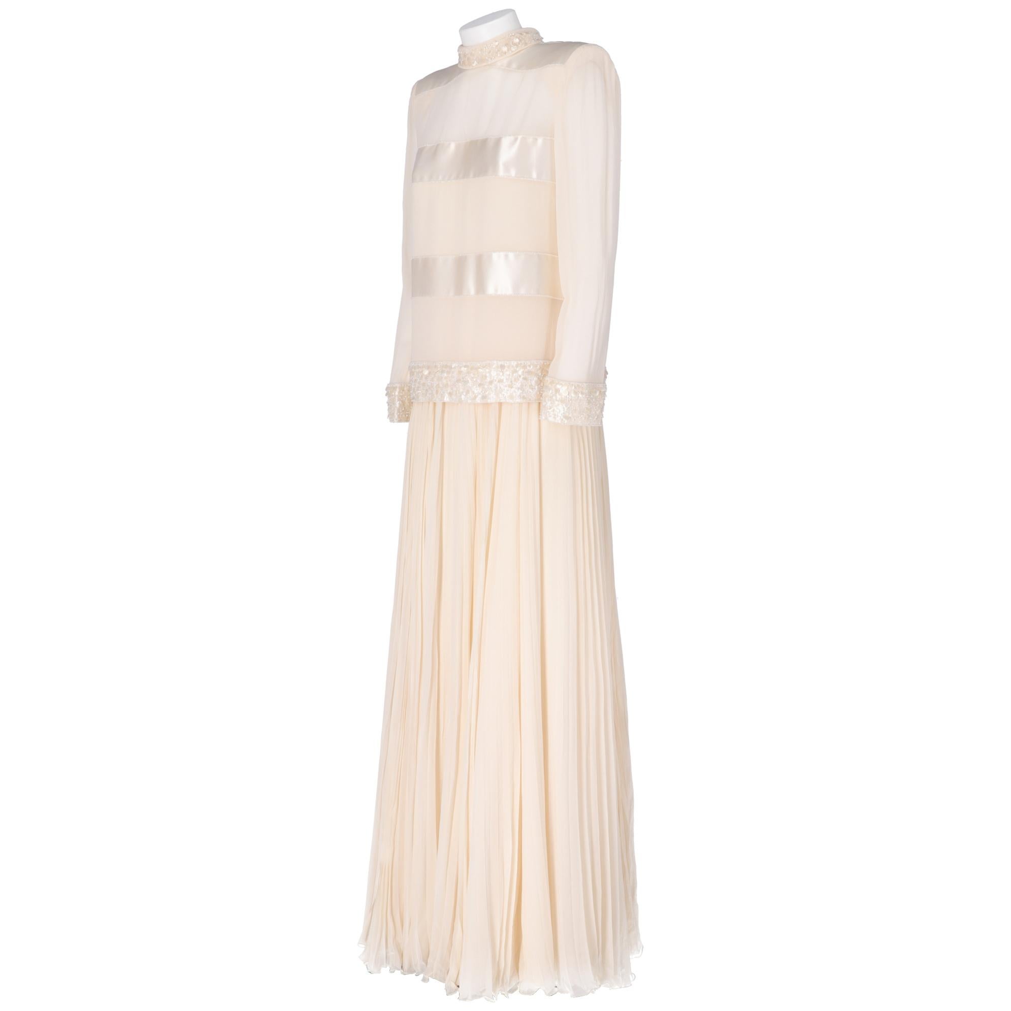 Tailored wedding dress in ivory-colored silk georgette, consisting of a two-ply pleated skirt with inner lining and collar with spikes on the back, padded straps, decorative bands in silk satin and beaded and sequined appliqués bow. Back zip