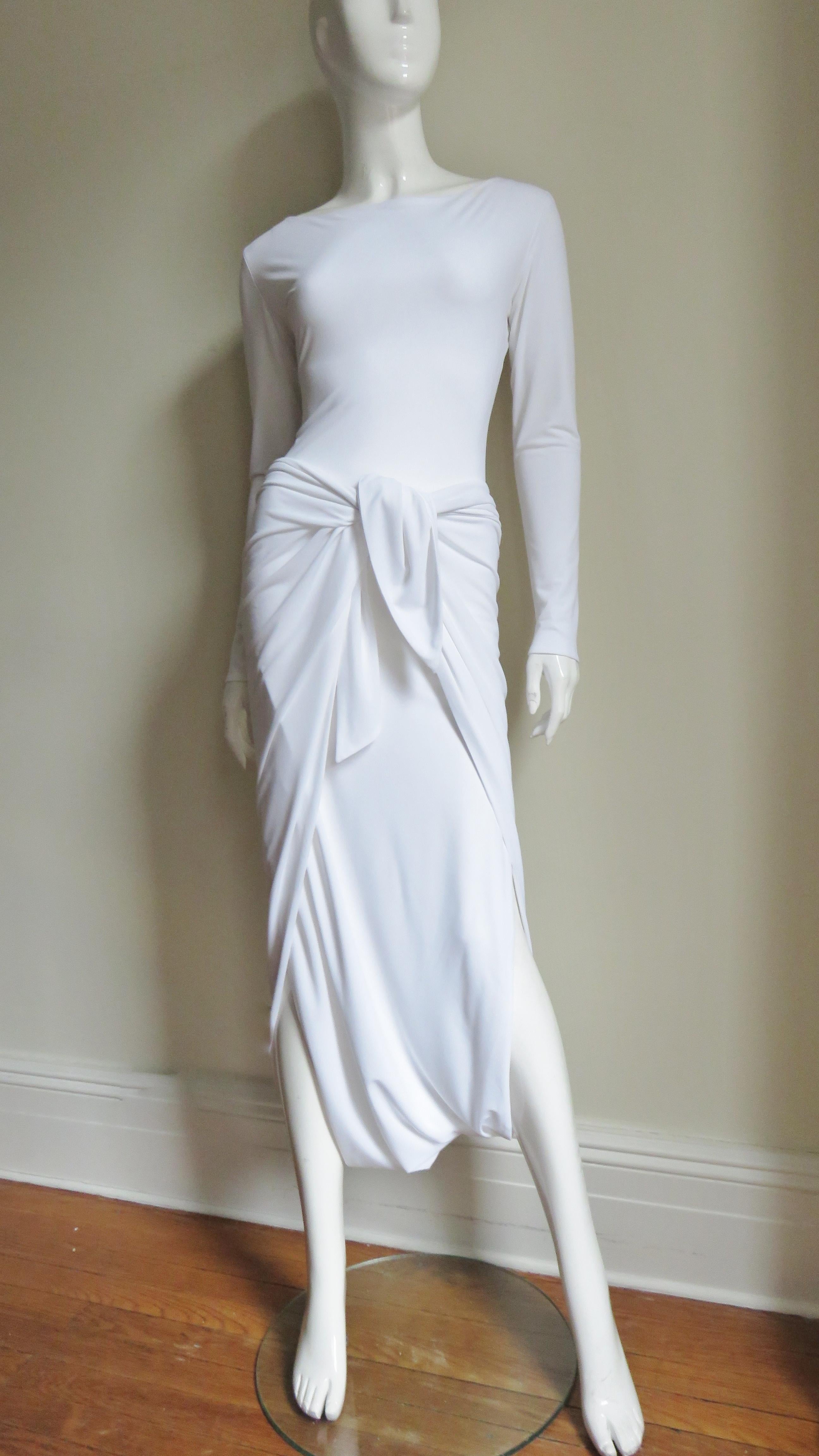 A fabulous unique white silk jersey dress from Norma Kamali.  It Has long sleeves, a bateau neckline and is semi fitted through the waist via a long front panel that drapes in front then between the legs wrapping at the back hips the corners tying