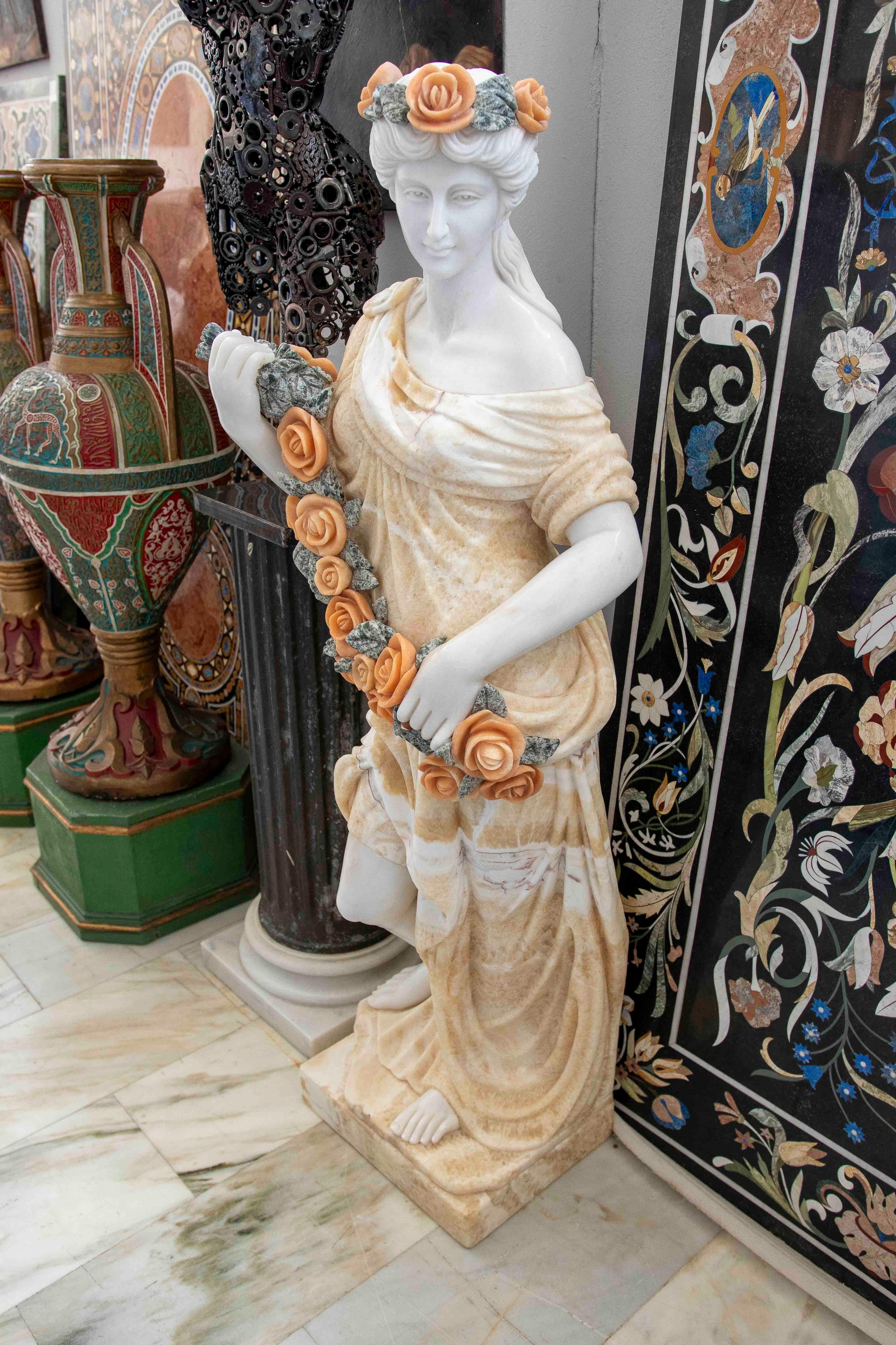 An exceptional pair of finely carved polychrome marble sculptures depicting allegorical female figures of Spring. Each figure appears standing, wearing a draped dress in warm tones, holding a garland of flowers sculpted in contrasting marble, with